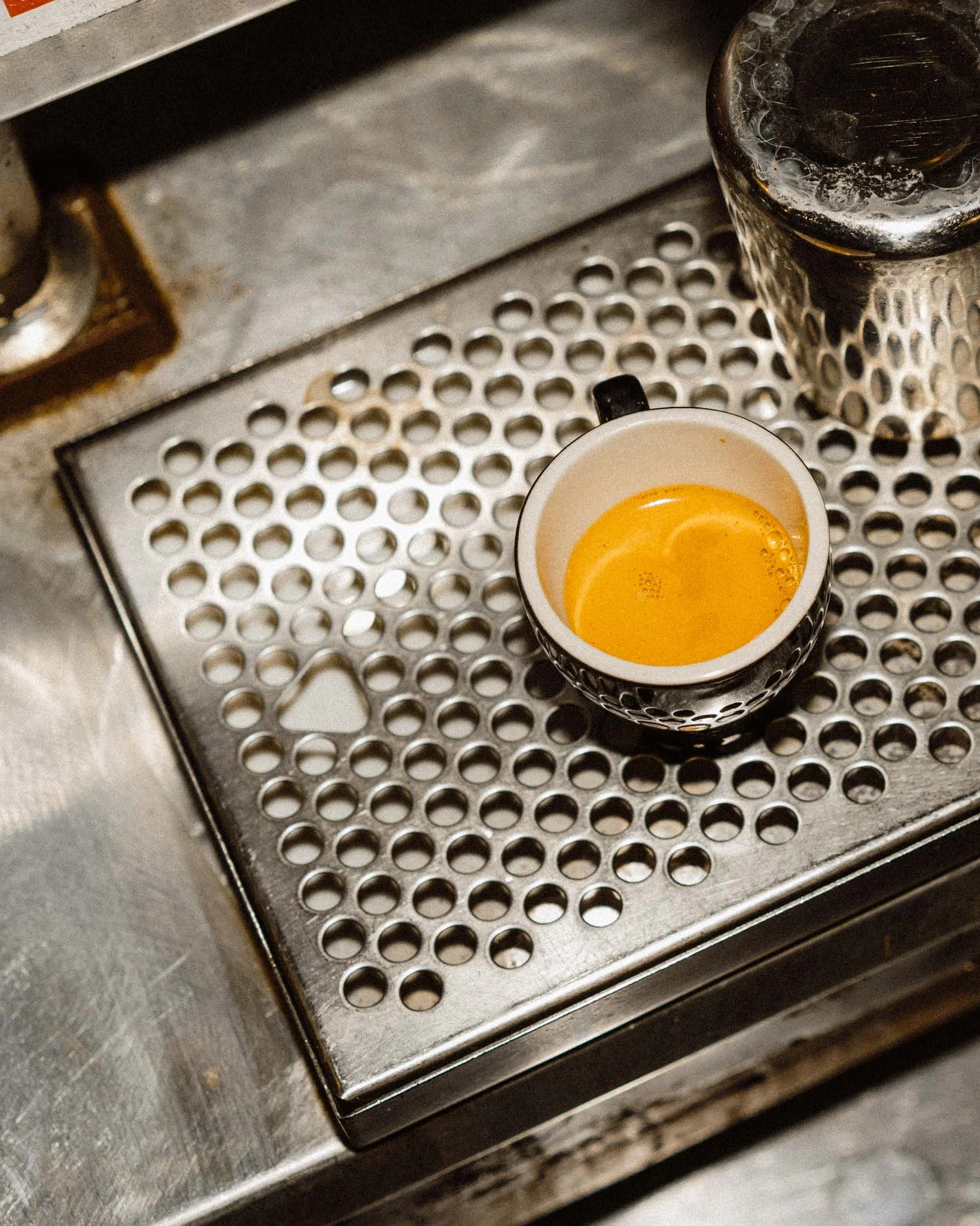 A cup of freshly brewed espresso on a metal drip tray with a glass container nearby.