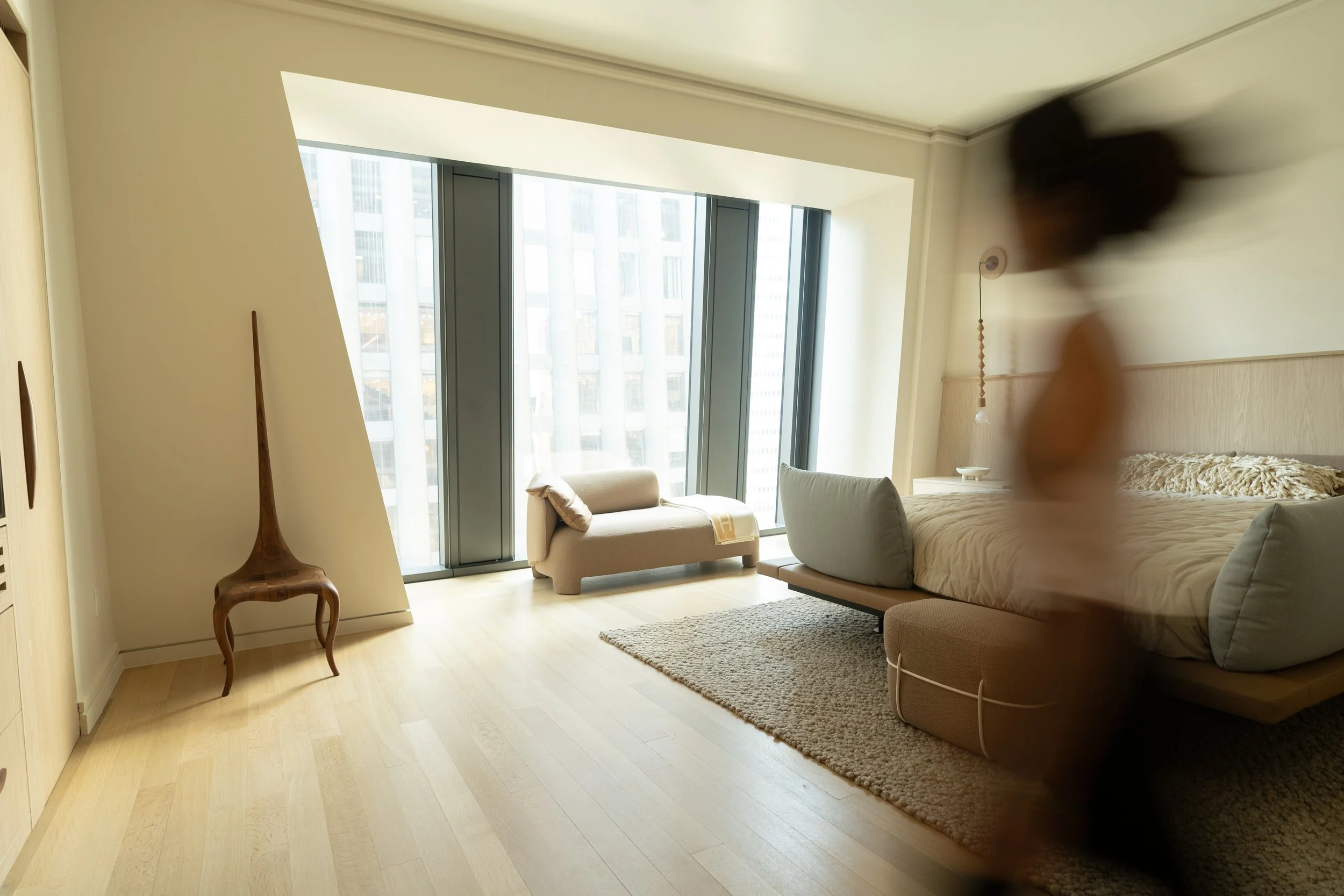 Modern bedroom with floor-to-ceiling windows, a beige bed with pillows and a small beige ottoman, a cozy loveseat near the window, a textured area rug, a wooden decorative sculpture, and a blurred silhouette of a person.