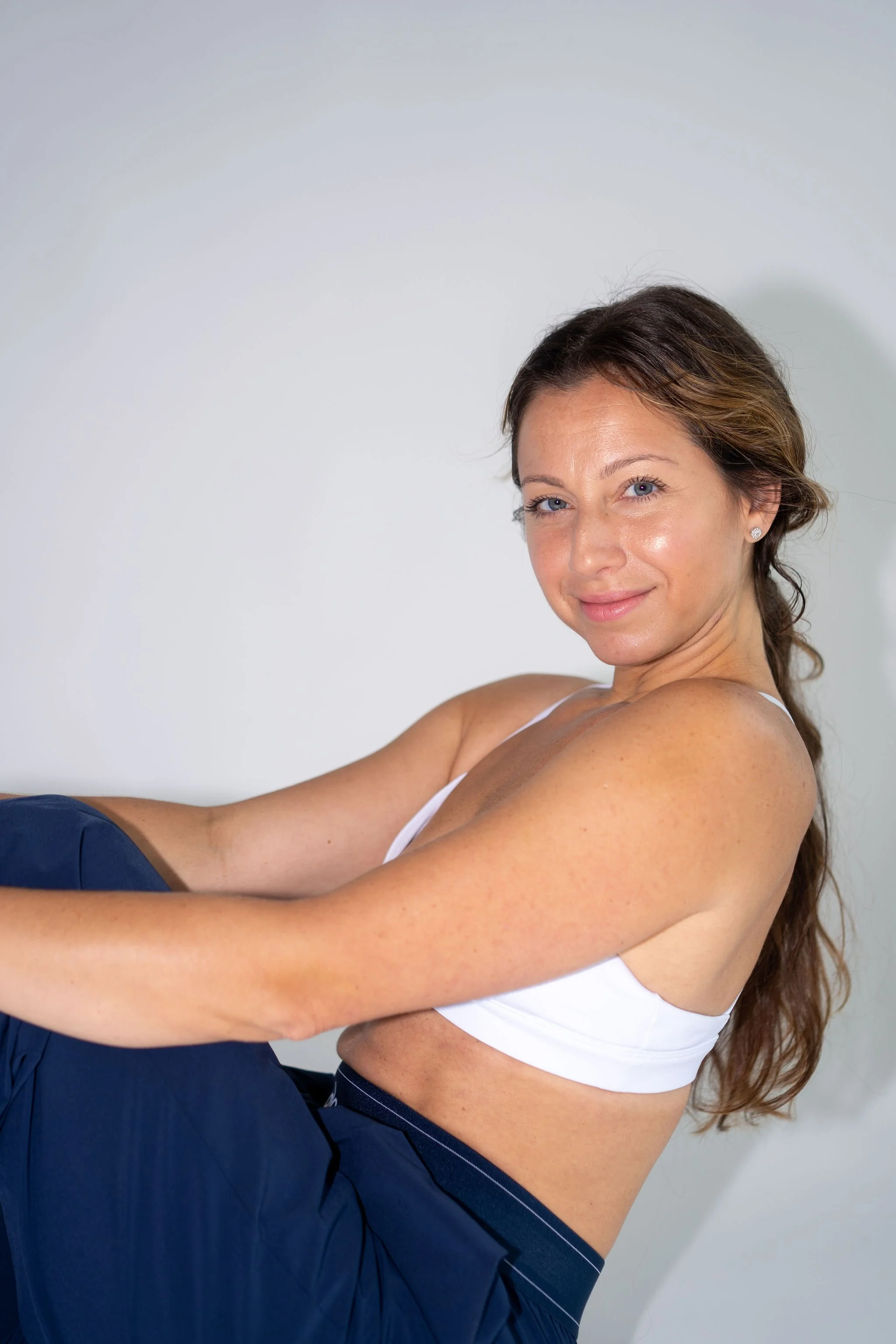 A woman with long brown hair and blue eyes, wearing a white tank top and navy pants, sitting against a plain white wall, smiling at the camera.