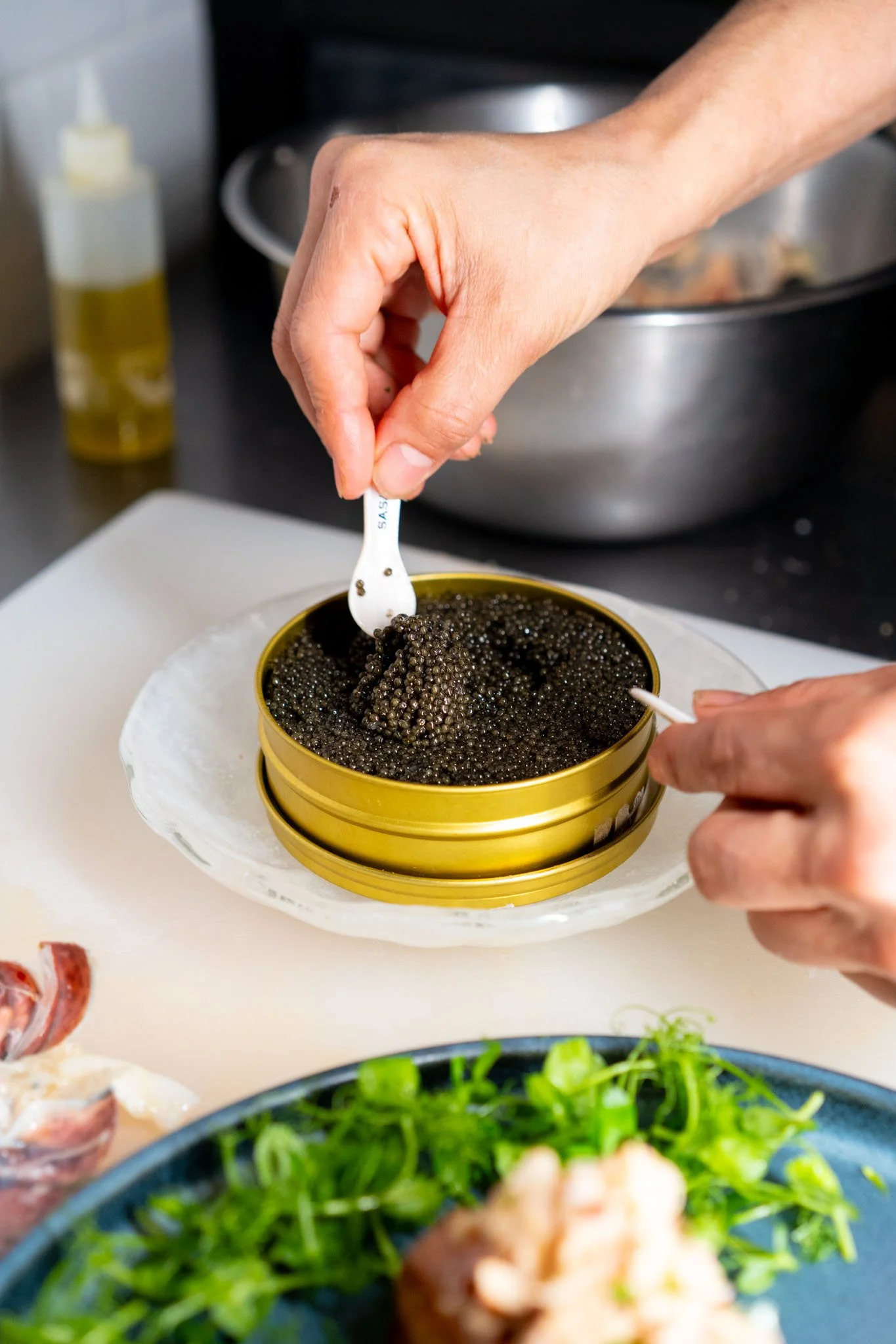 A person using a tiny spoon to scoop black caviar from a gold tin container on a white plate, part of a food preparation scene.