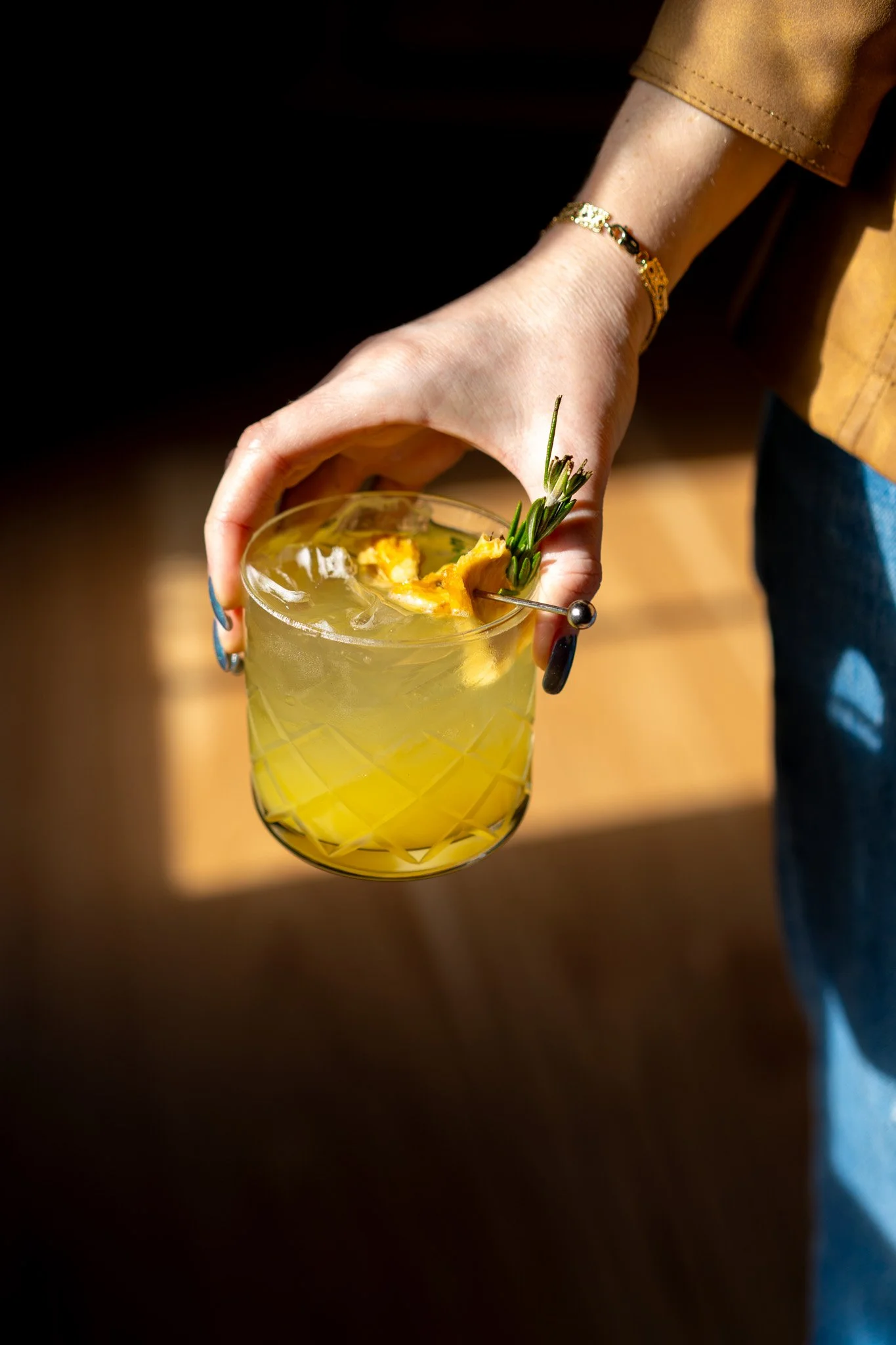 Hand holding a glass with a yellow cocktail garnished with herbs and pineapple with a decorative pick.