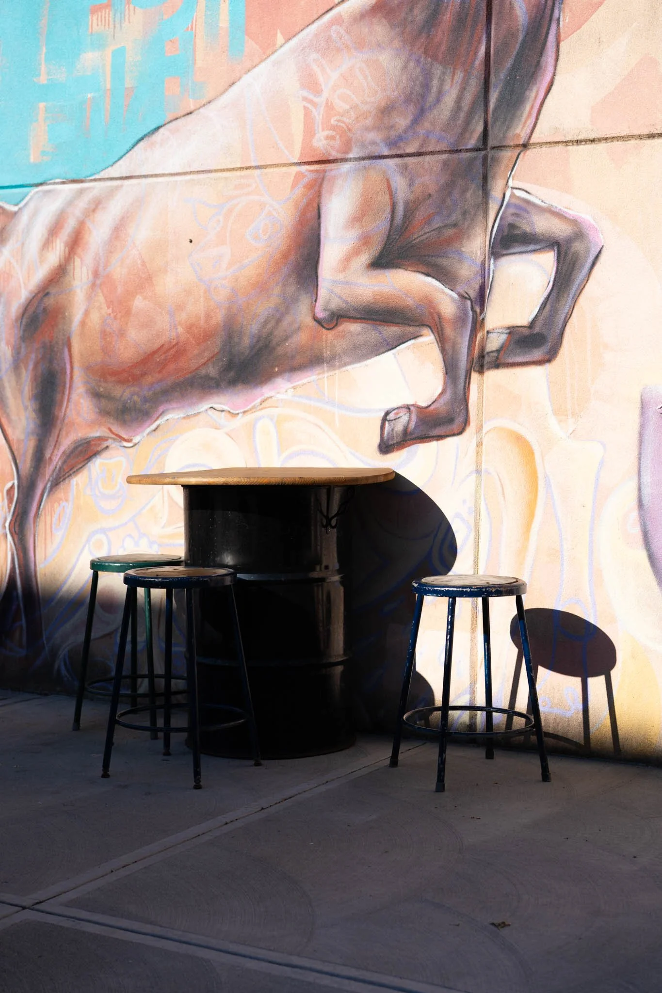 Outdoor seating area with a wooden table and two metal stools in front of a colorful graffiti wall featuring a stylized human figure.