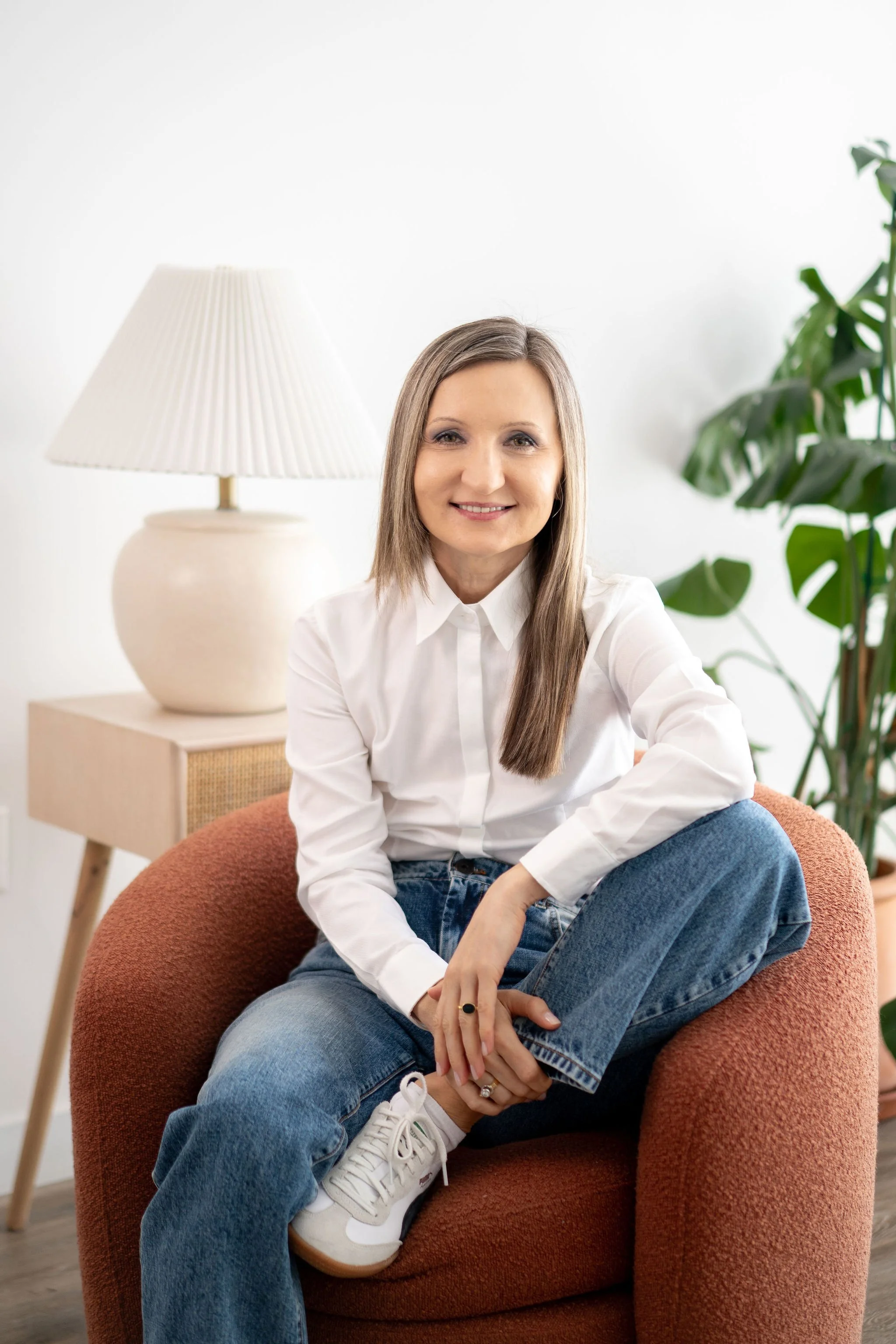 A woman with long brown hair, wearing a white blouse and blue jeans, sitting cross-legged on a rust-colored armchair in a bright, modern living room with a white wall, a table lamp, and green plants in the background.