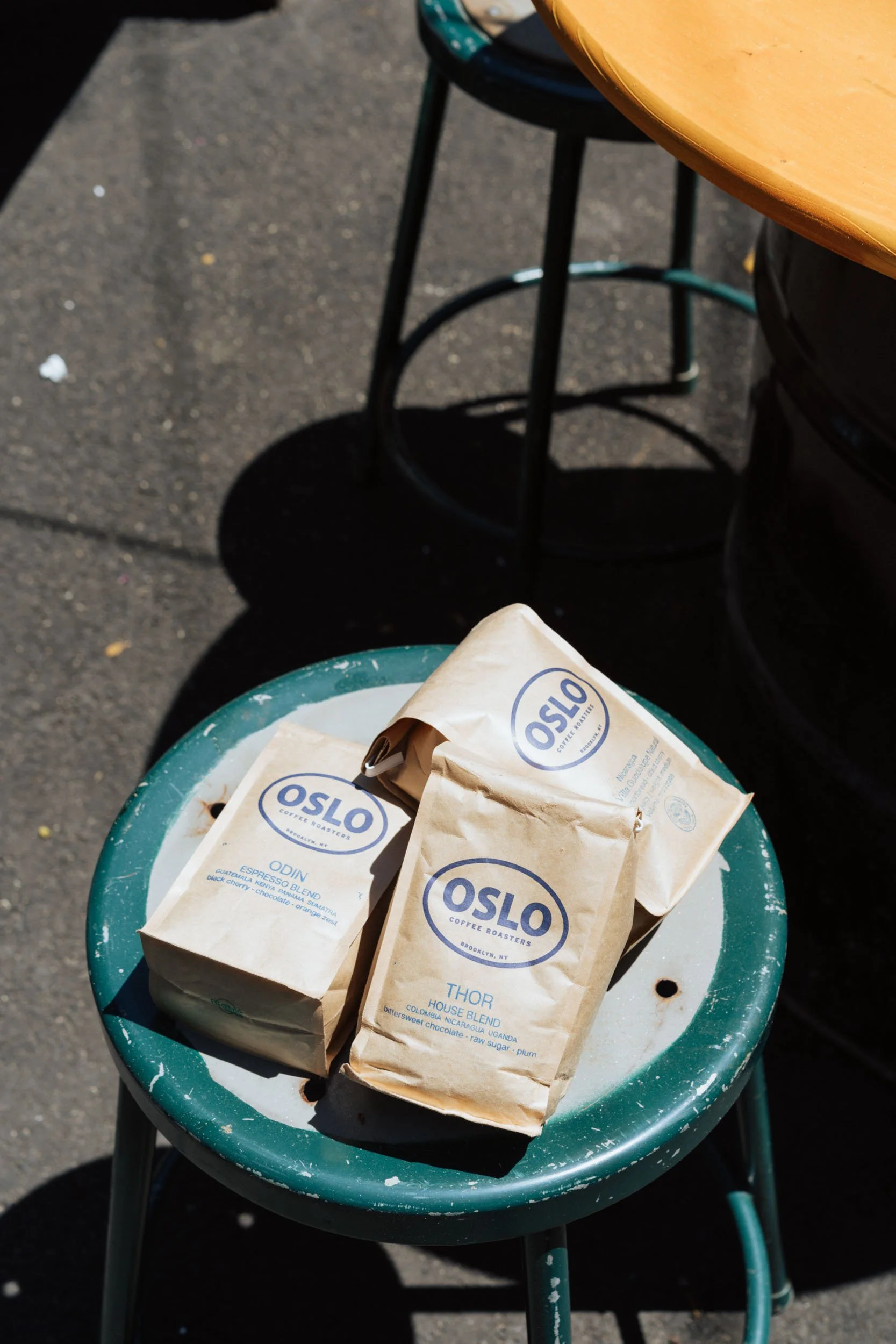 Three bags of Oslo coffee placed on a green, slightly worn outdoor stool, with a yellow table partially visible at the top right, and a concrete ground with shadows in the background.