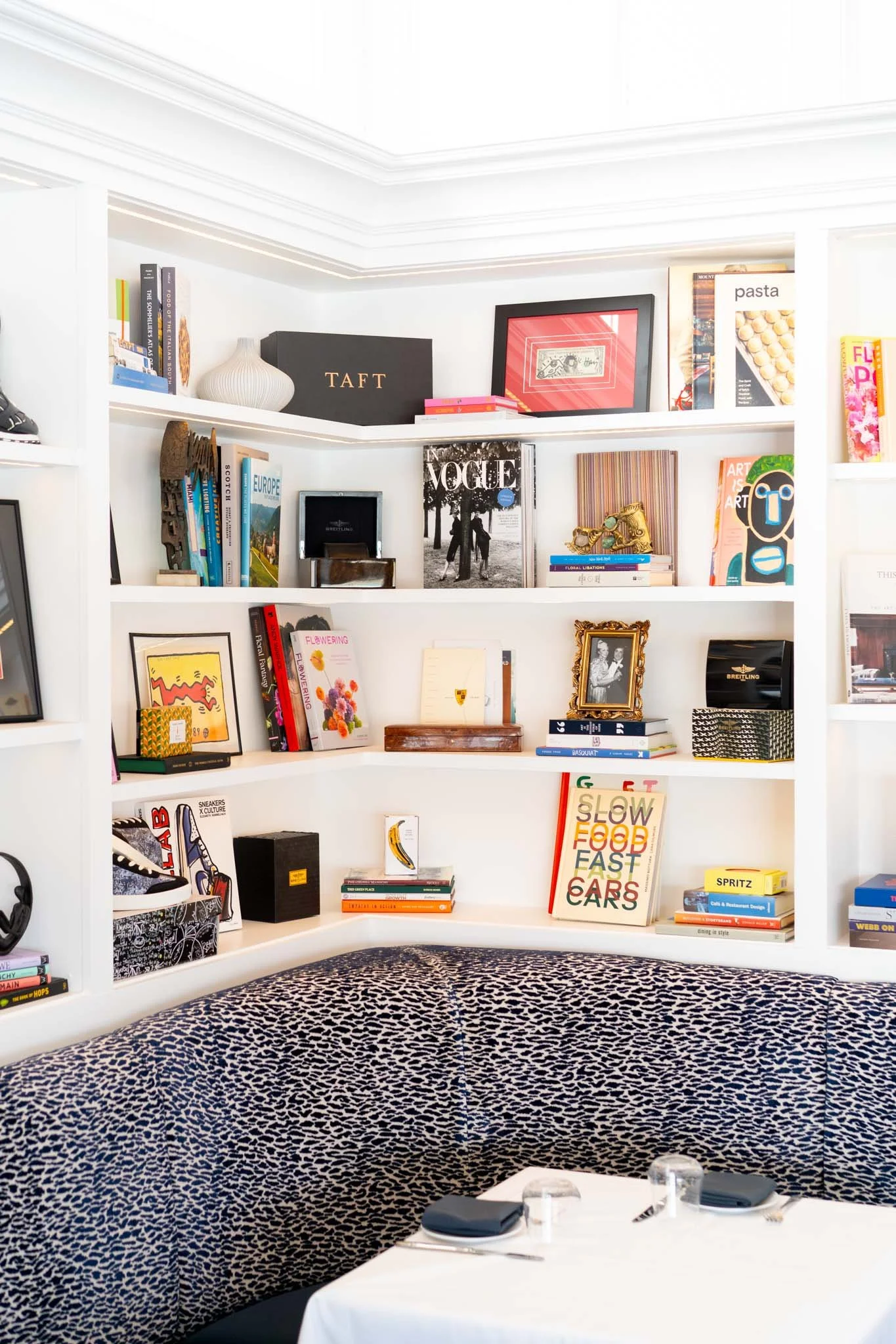 White built-in bookshelf filled with books, photo frames, decorative items, and awards, with a curved, animal print upholstery booth and a table with place settings in the foreground.
