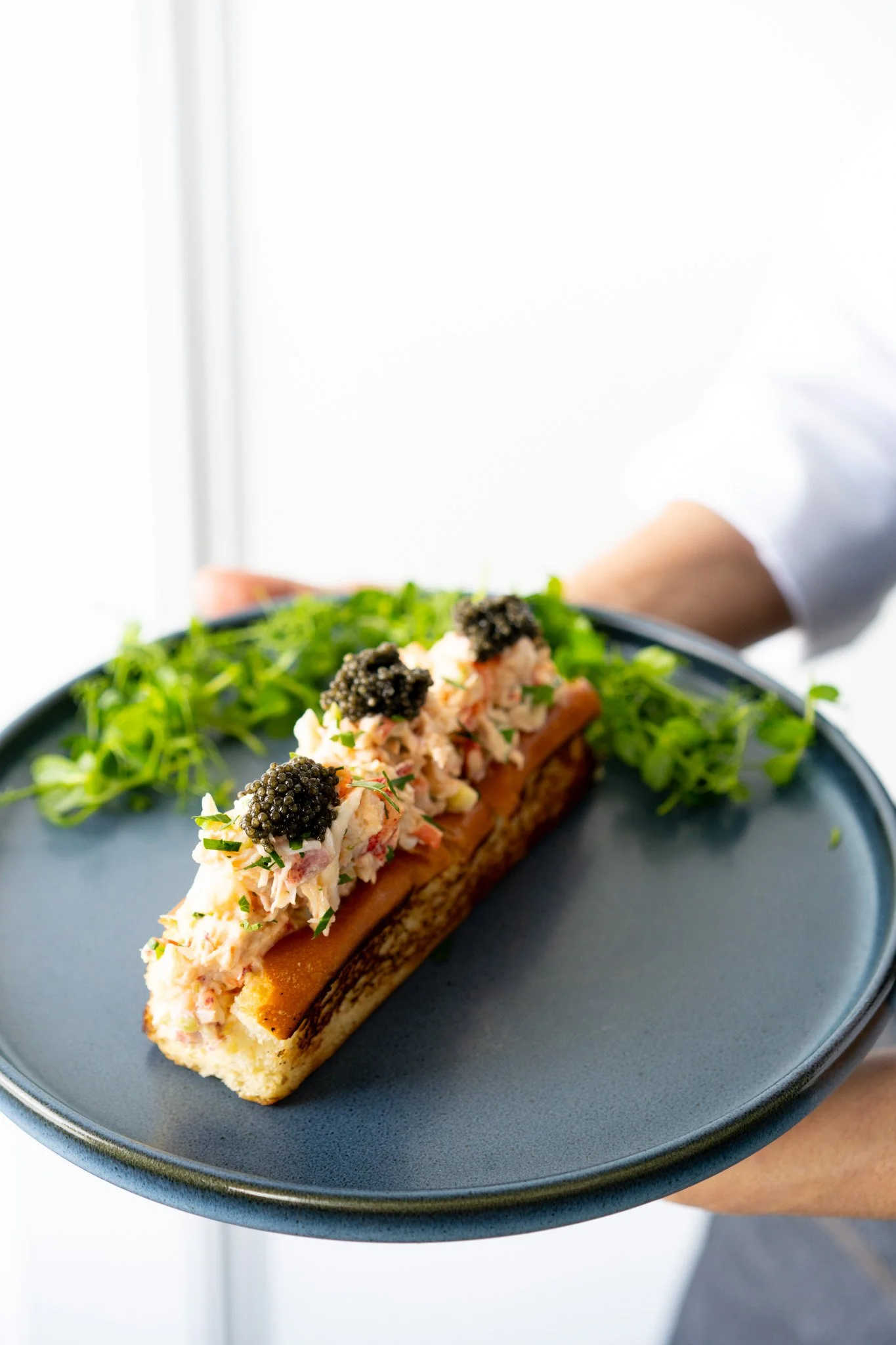 Lobster roll with caviar topping on a navy plate with greens in the background.