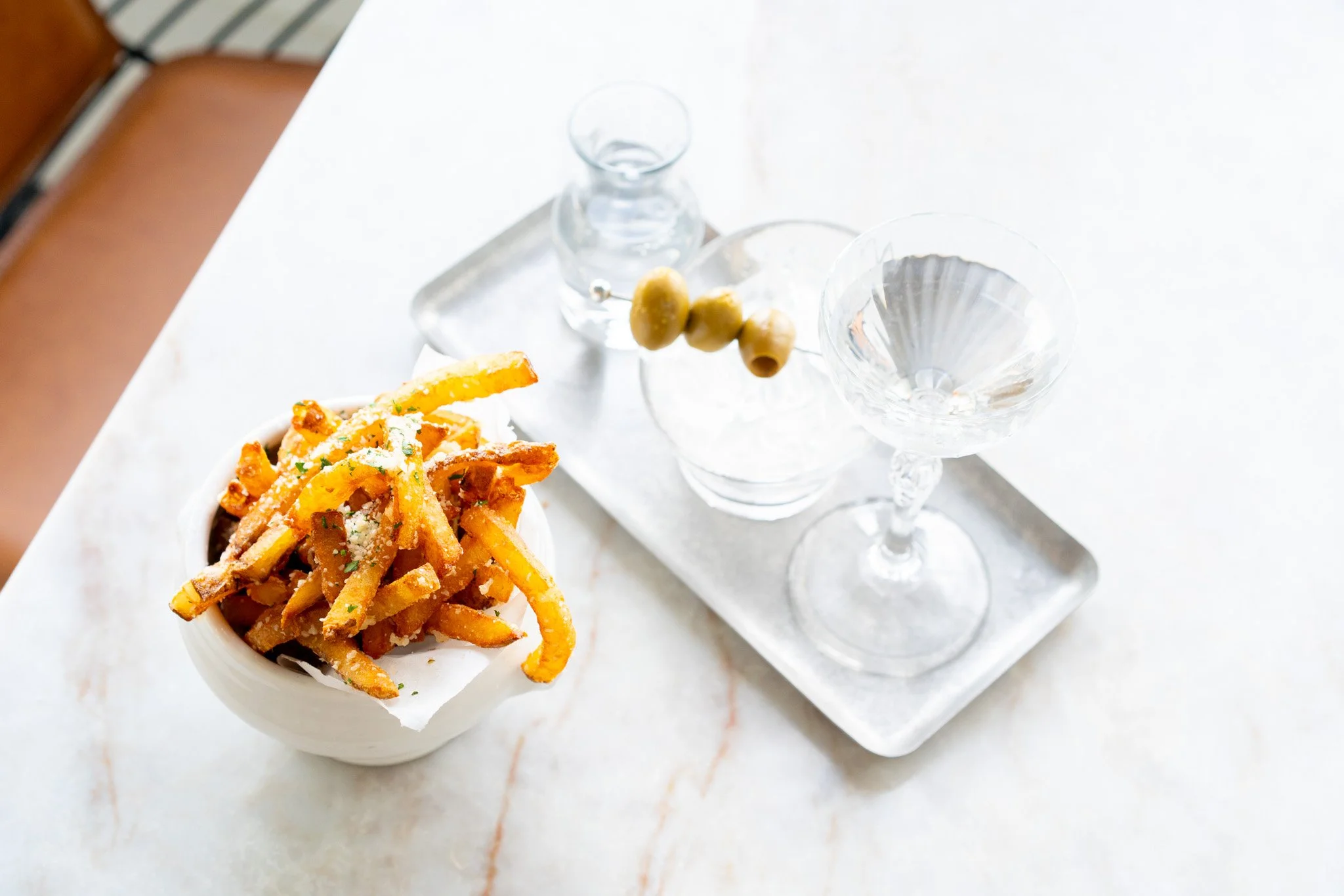 A white bowl of French fries topped with grated cheese on a white marble table, with a tray holding three empty cocktail glasses and a small glass water pitcher.