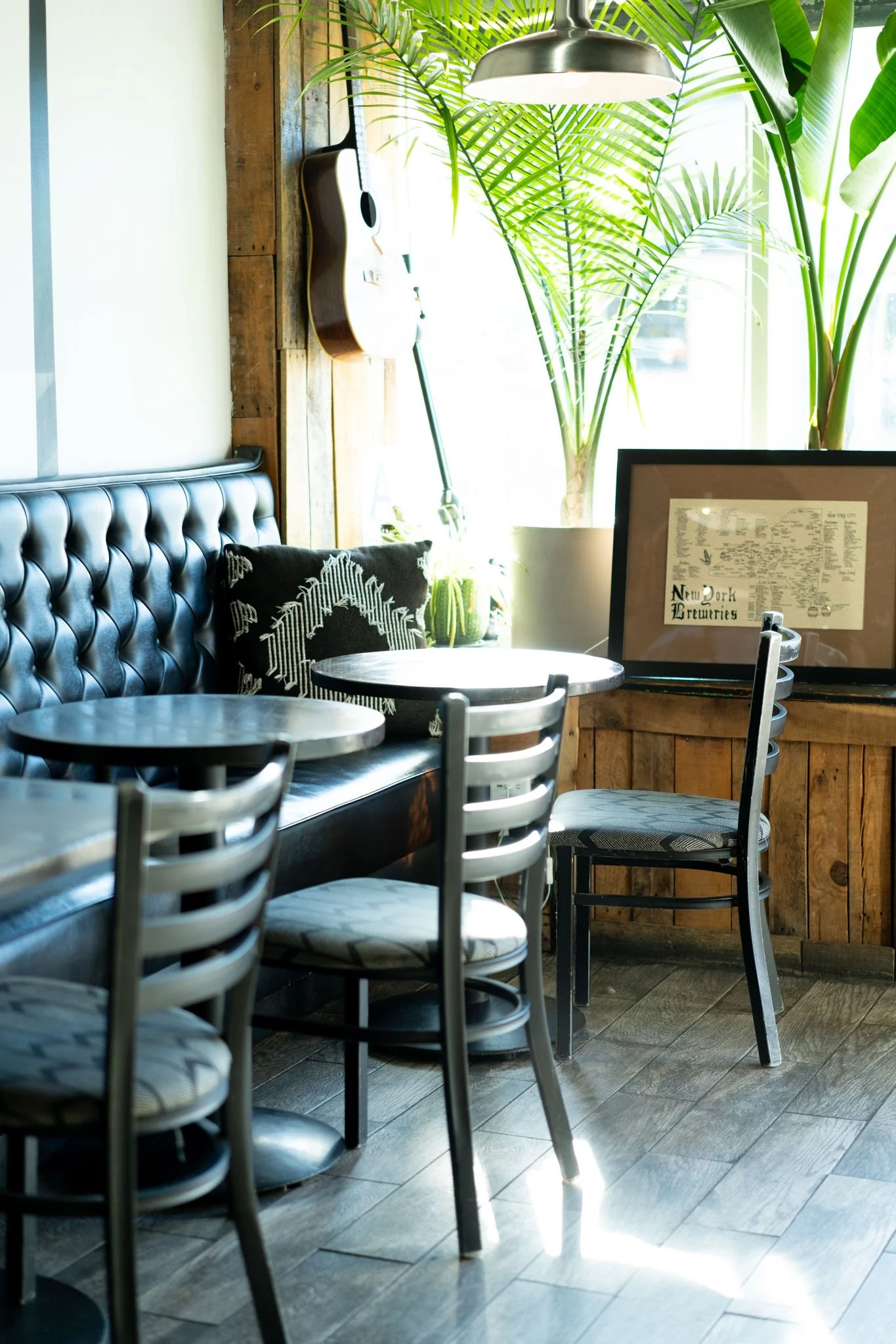 A cozy cafe corner with black leather tufted banquette seating, wooden panel walls, black tables and chairs, large green potted plants, a wall-mounted guitar, framed artwork, and natural sunlight.