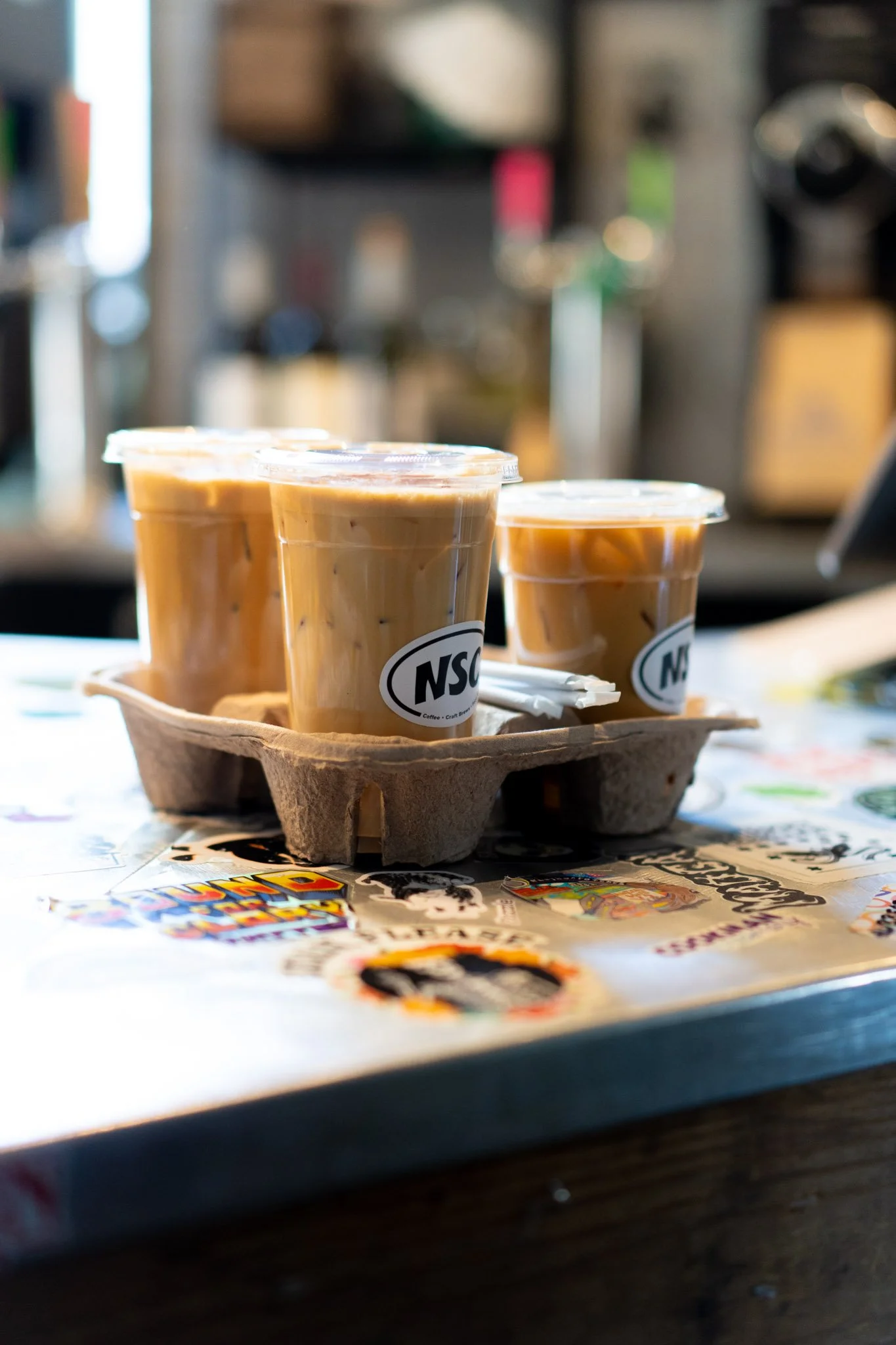 Three cups of iced coffee in plastic cups with lids, on a tray, placed on a table with colorful stickers.