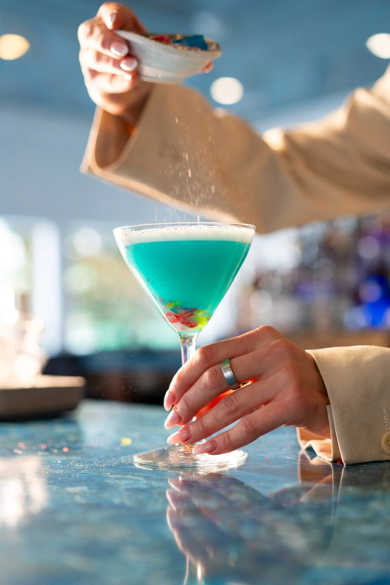 A person pouring a blue cocktail with colorful toppings into a martini glass at a bar.