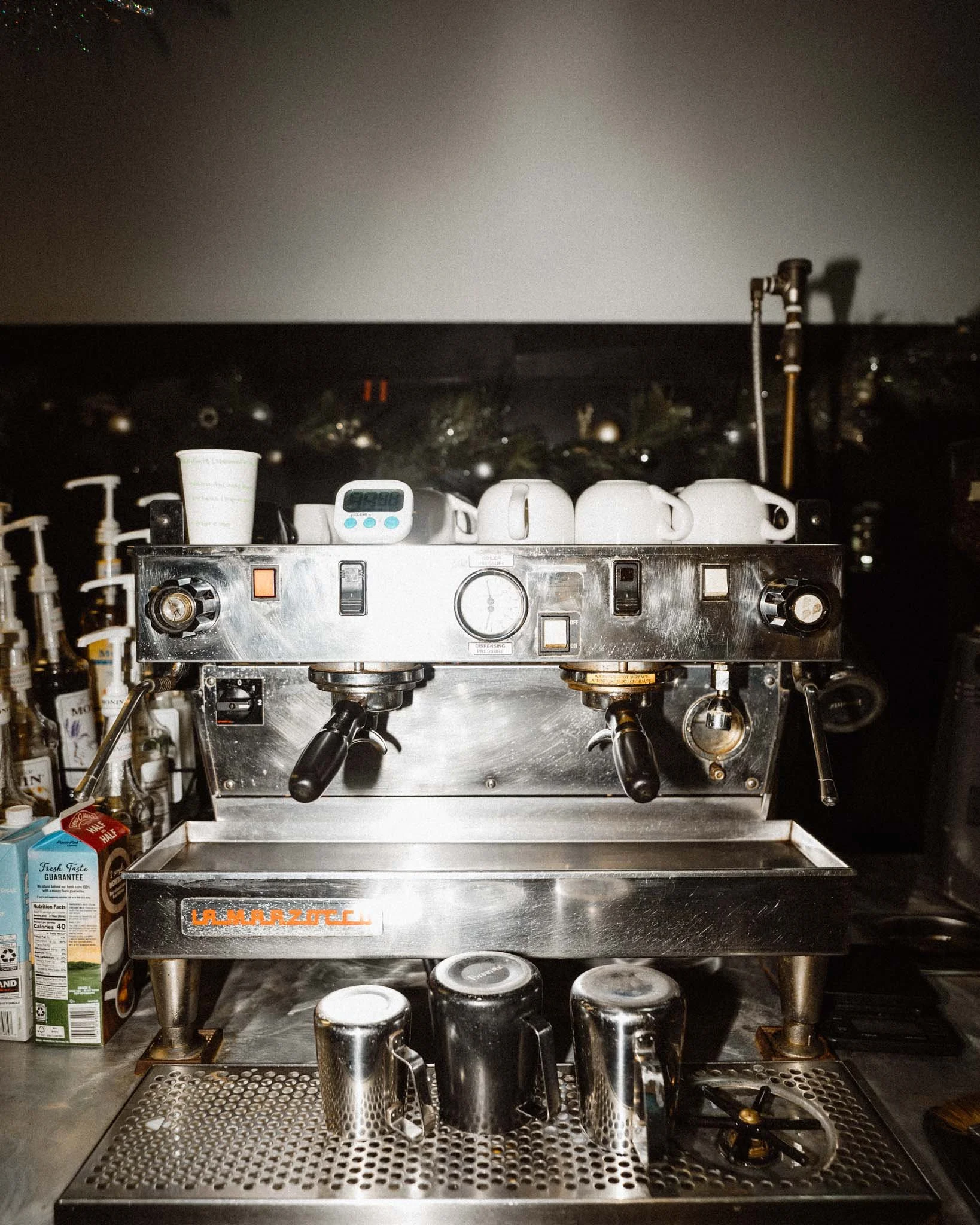 A commercial espresso machine with four white cups on top, a small digital timer, and various controls, located on a metal counter with coffee-related supplies and a row of cups at the front.