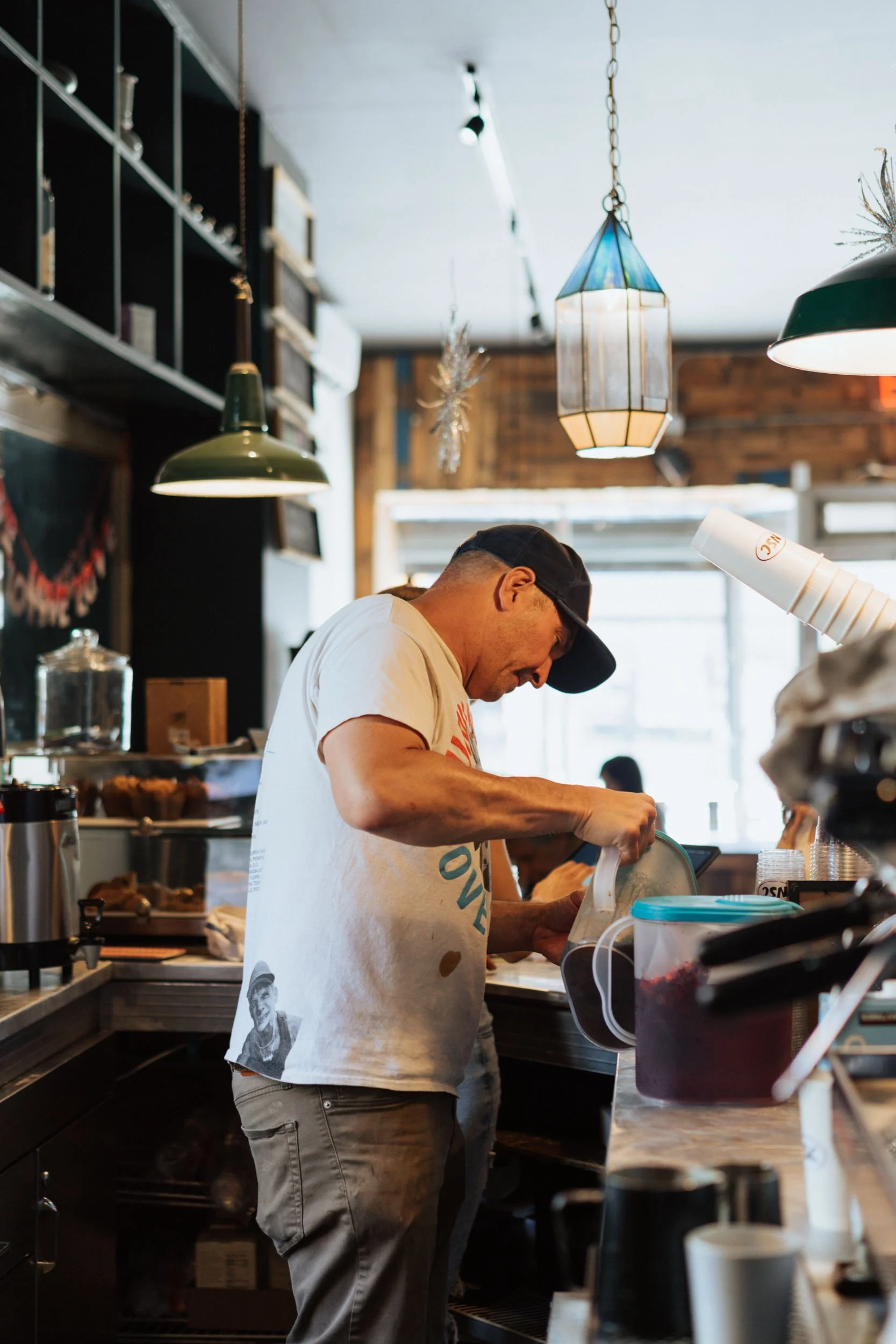 A man in a white T-shirt and baseball cap working behind a coffee shop counter, pouring something into a container.