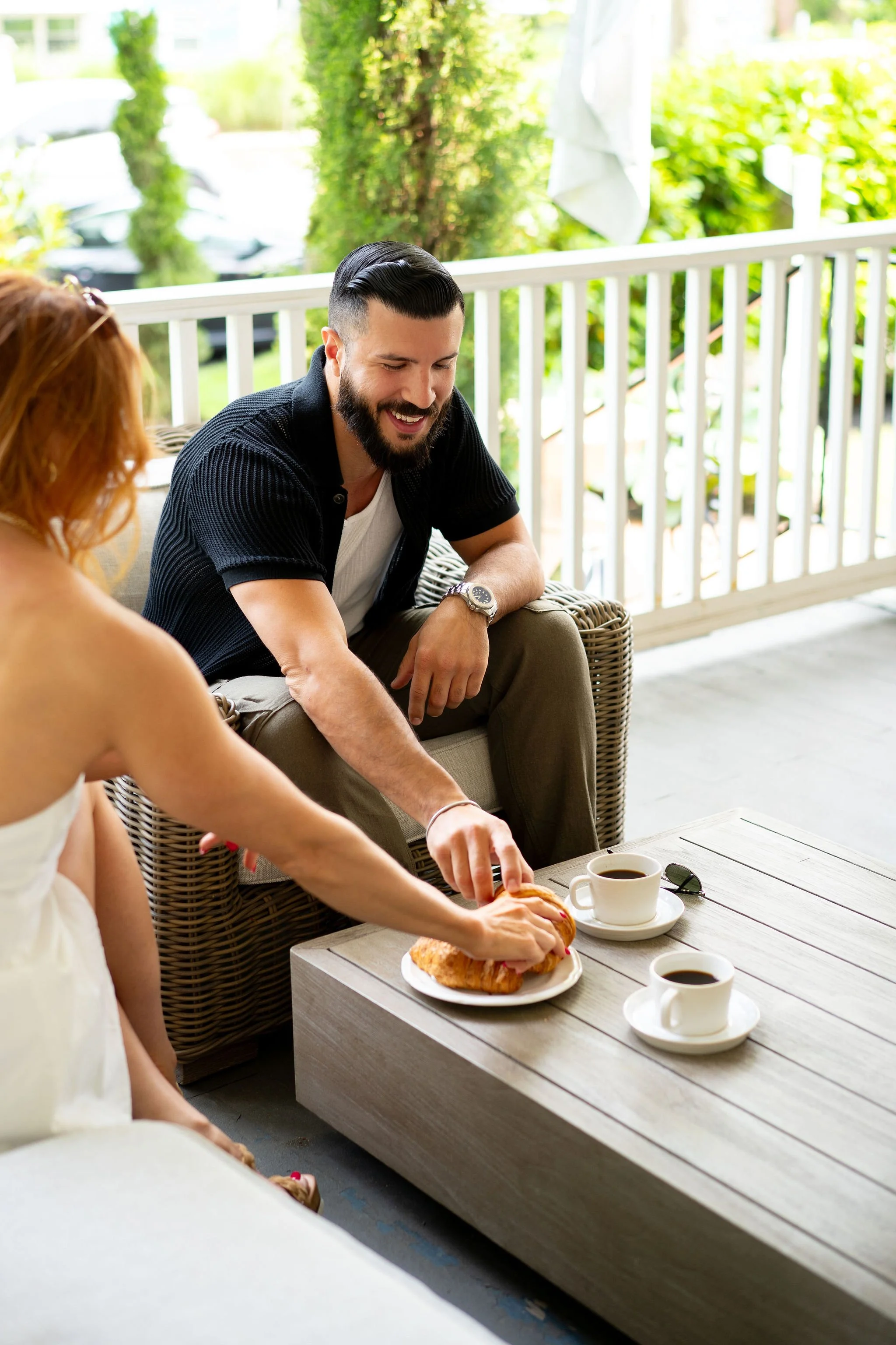 A man and woman sitting on a porch, sharing a moment over coffee and a pastry. The man is smiling, reaching for the pastry, while the woman is in a white dress, leaning forward. Two cups of coffee and a pastry are on the table.