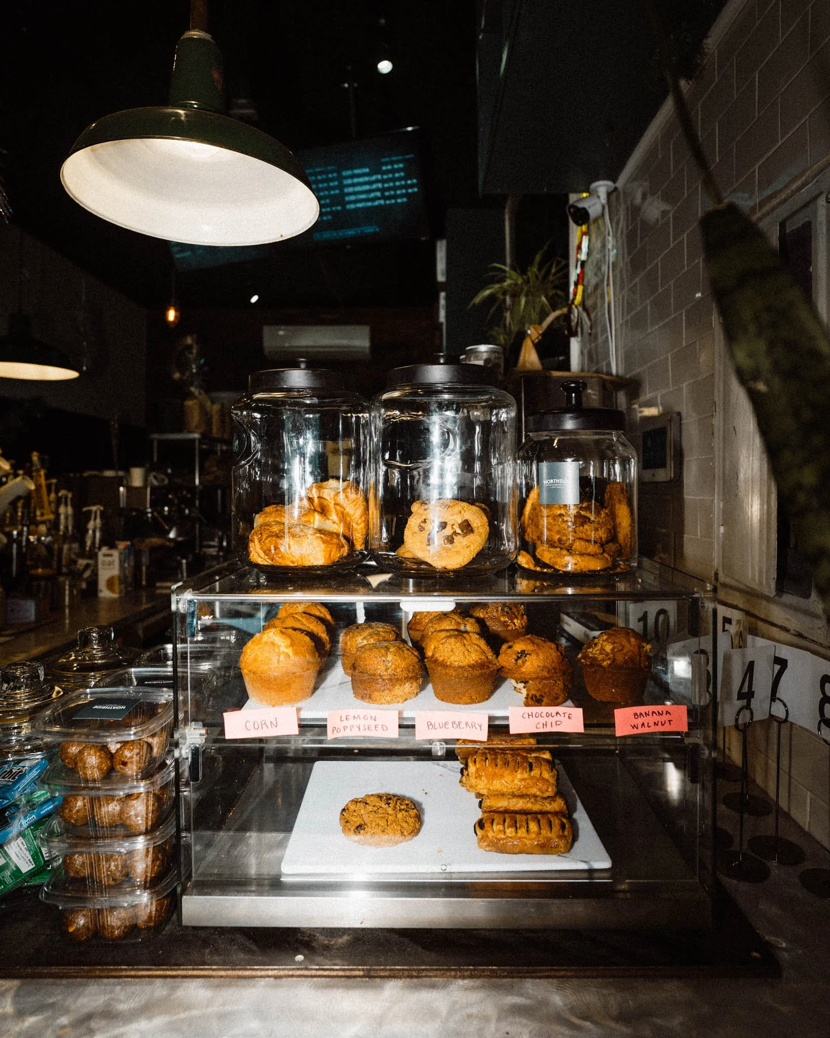 Display case with cookies and muffins at a bakery or cafe, labeled with flavors like corn, lemon poppyseed, blueberry, chocolate chip, and banana walnut.