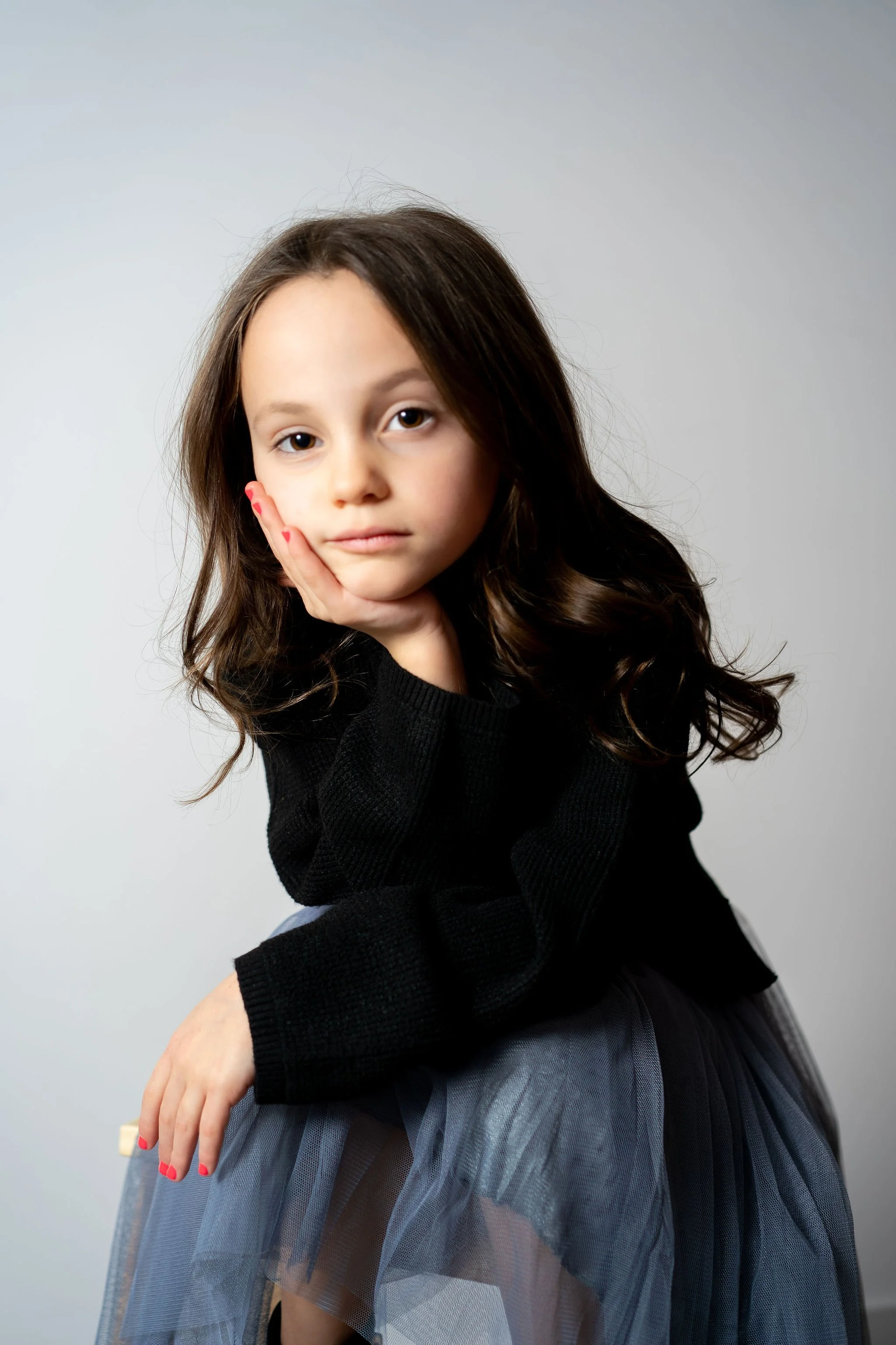 A young girl with brown hair and a black sweater sitting against a gray background, posing with her head resting on her hand.