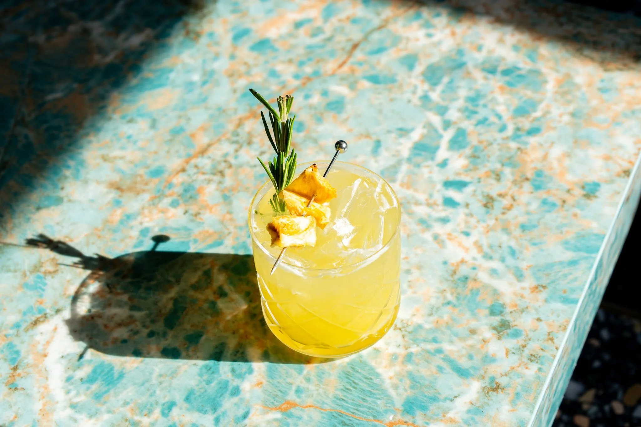 Yellow cocktail with ice, garnished with pineapple chunks and a sprig of rosemary, sitting on a colorful marble surface with a shadow cast by the drink.