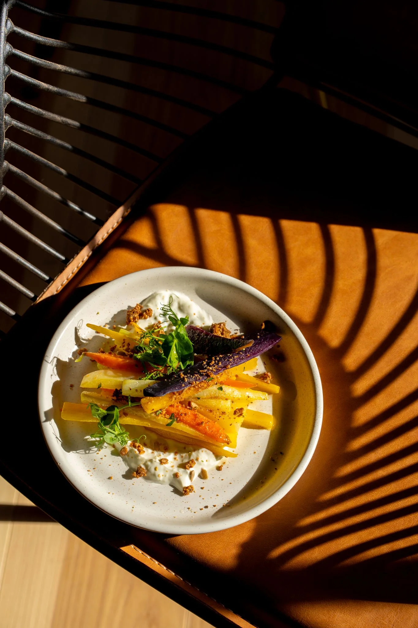 A bowl of roasted vegetables with whipped cream, garnished with microgreens, served on a white dish. The bowl is placed on a orange leather surface with shadows cast from a slatted surface.