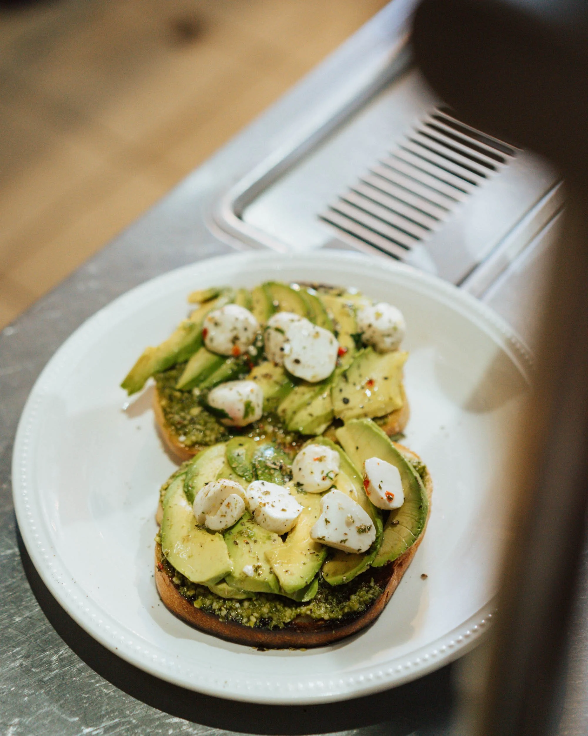 Two slices of avocado toast topped with mozzarella cheese on a white plate, placed on a stainless steel surface.