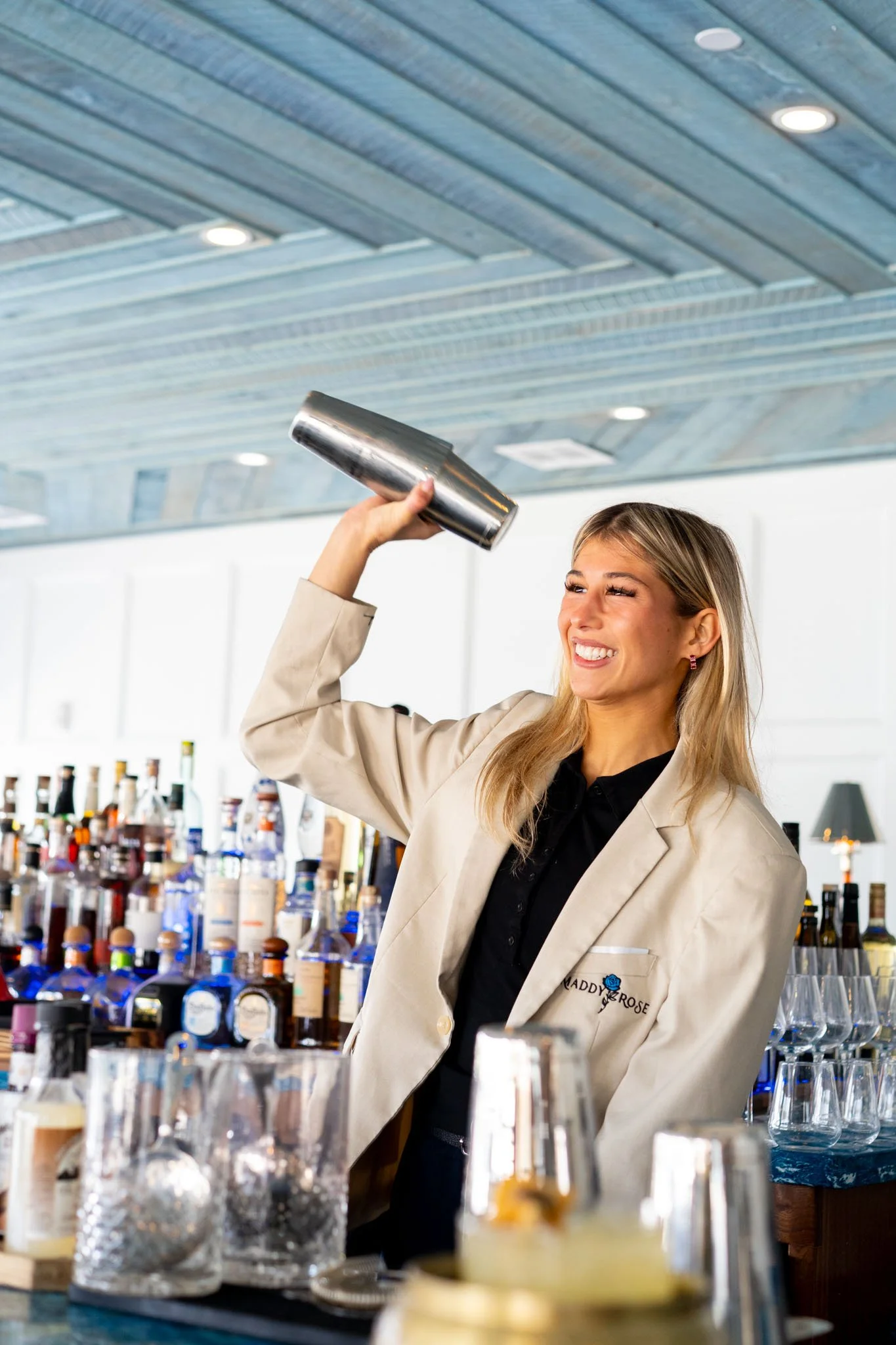 A smiling woman shaking a cocktail shaker behind a bar with bottles and glasses visible in the background.