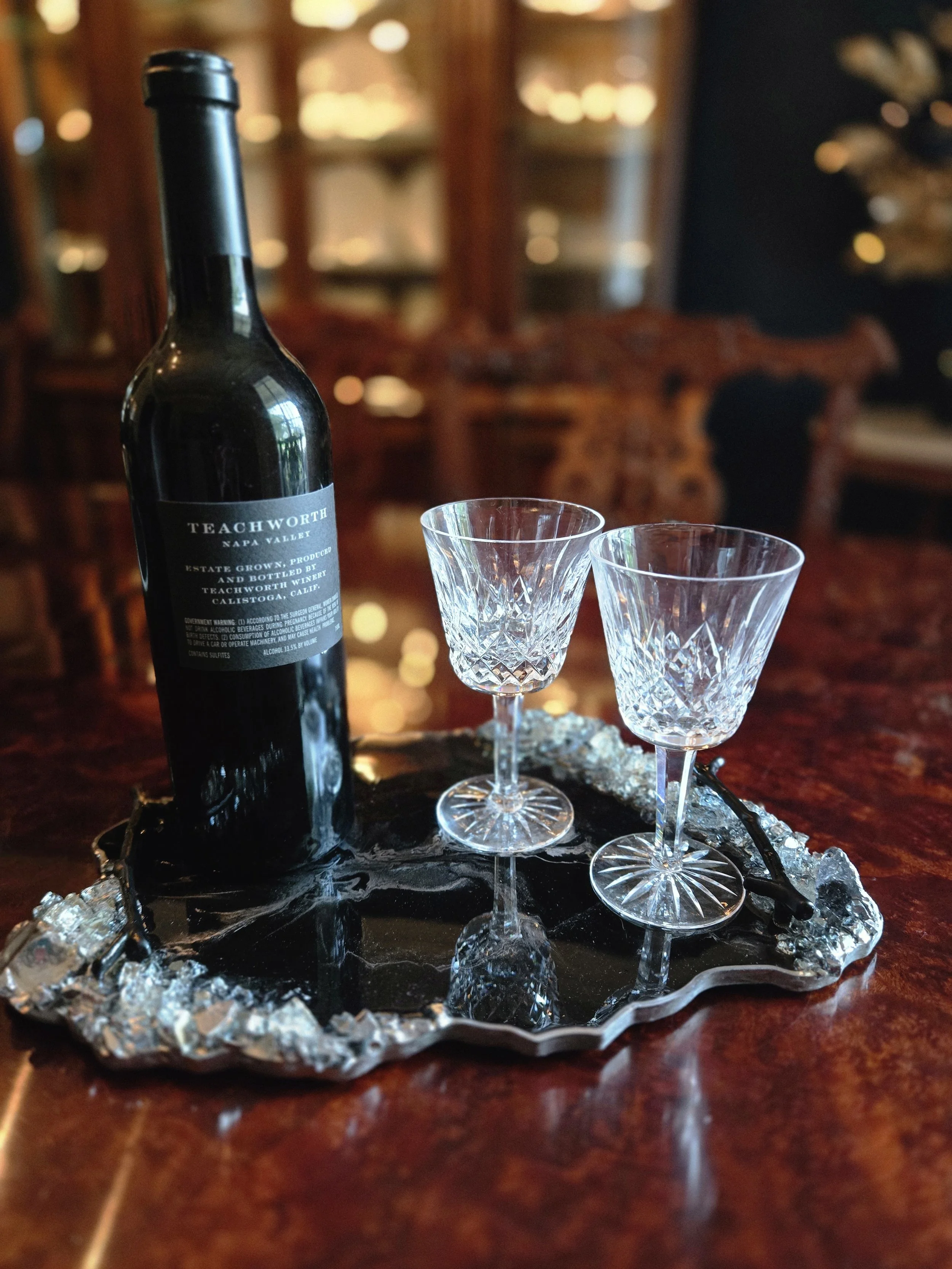 A bottle of Teachworth Napa Valley wine and two stemware glasses on a decorative serving tray with crystals, all placed on a wooden table with a blurred background of a wooden cabinet or shelves.