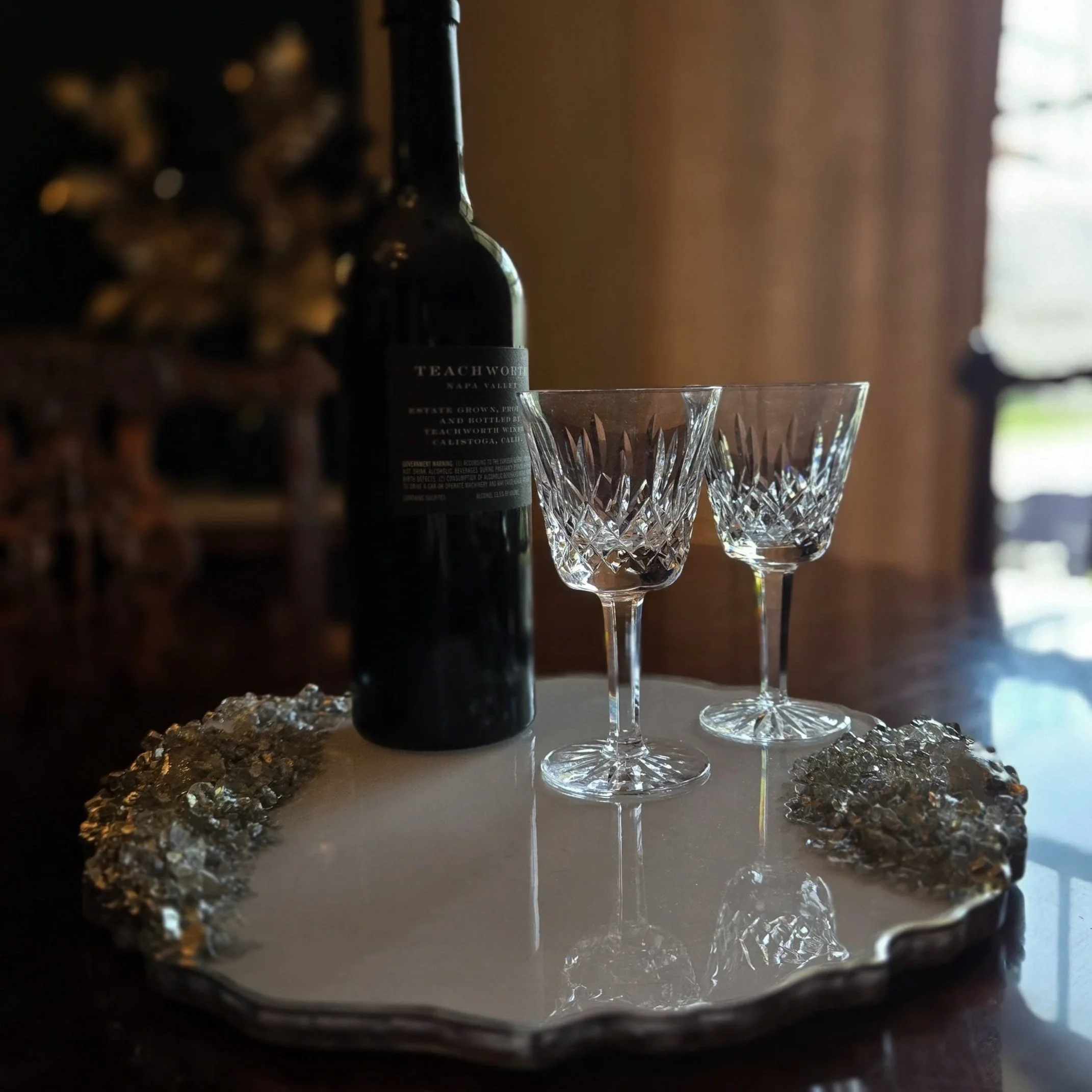 Close-up view of crystal-clear glasses with intricate patterns, placed on a reflective surface, with a dark wine bottle partially visible in the background. The scene is set indoors, illuminated by soft natural light coming from a window.