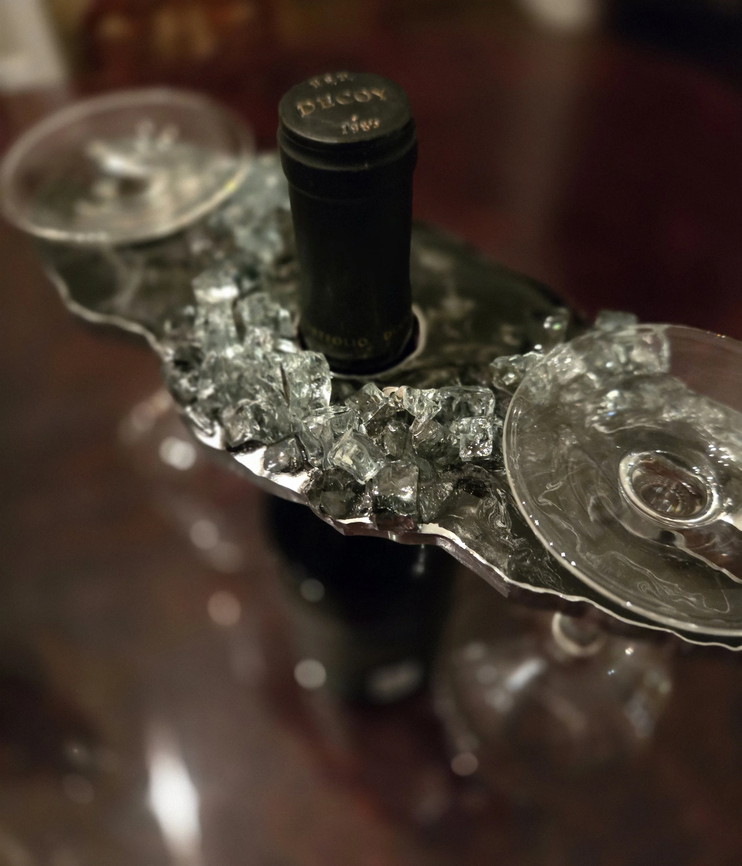 Close-up of a bottle cap with the words "ALCOHOL" and "187" on it, surrounded by ice cubes and part of a glass, with a blurred background.