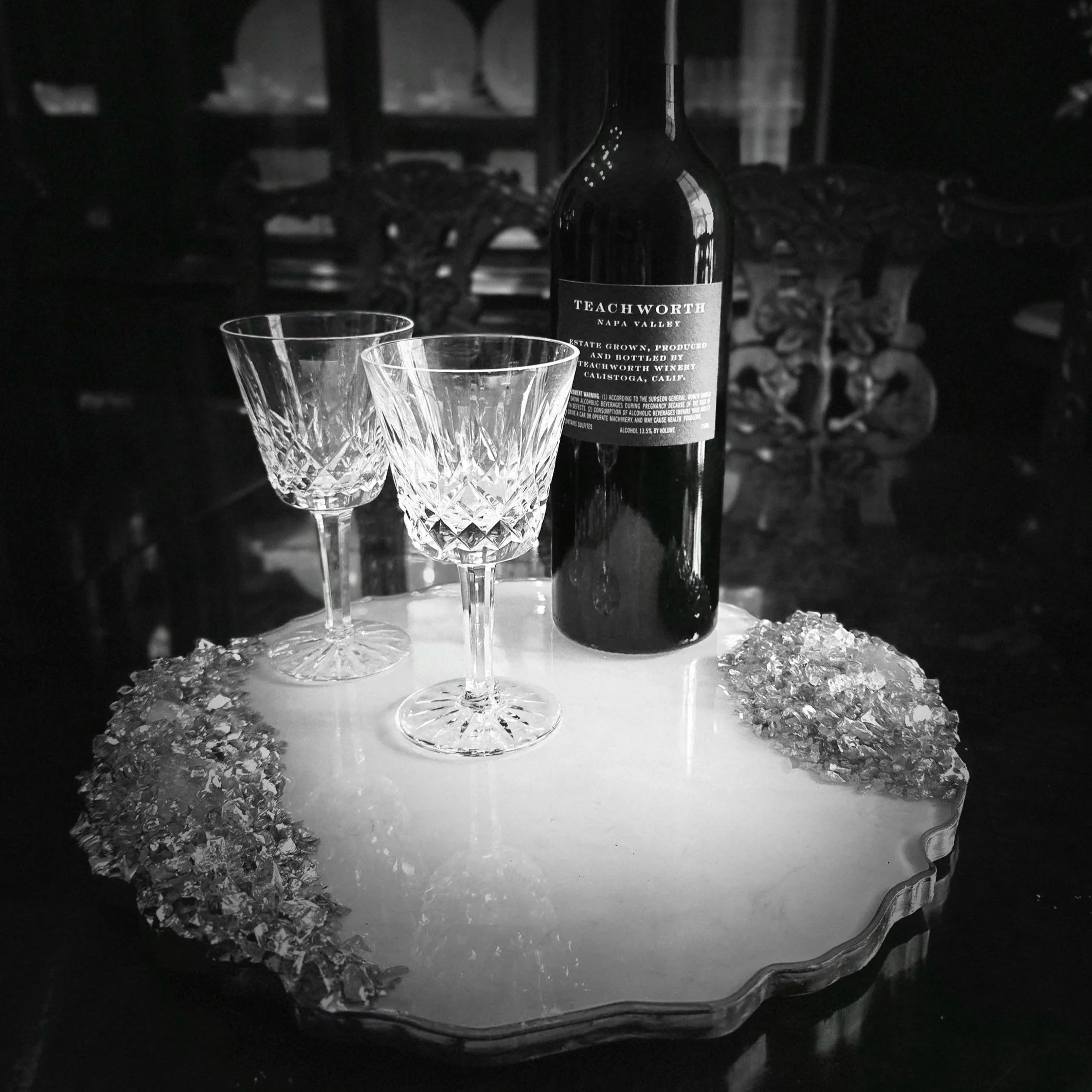 A bottle of Teachworth Napa Valley wine and two crystal wine glasses on a reflective surface with scattered ice, set against a dark background.