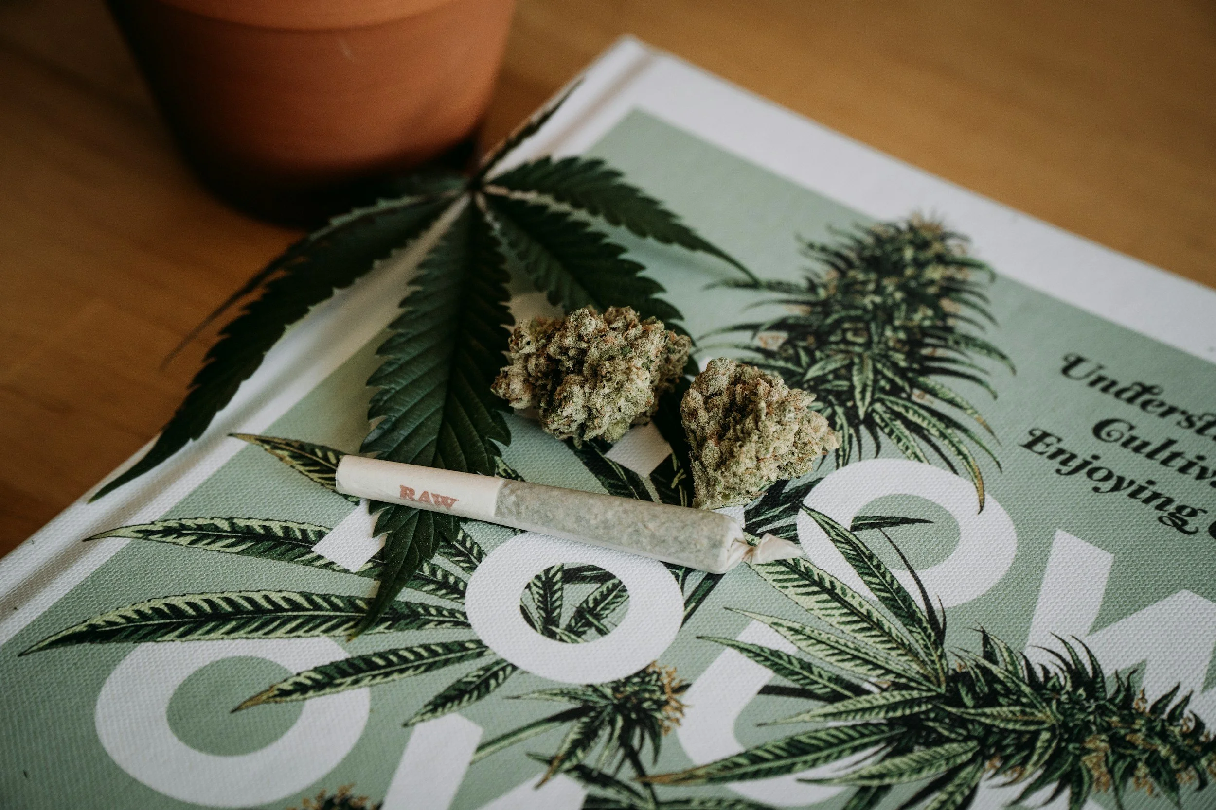 Cannabis buds, a marijuana leaf, and a joint placed on a magazine with cannabis-themed artwork and text, on a wooden surface.