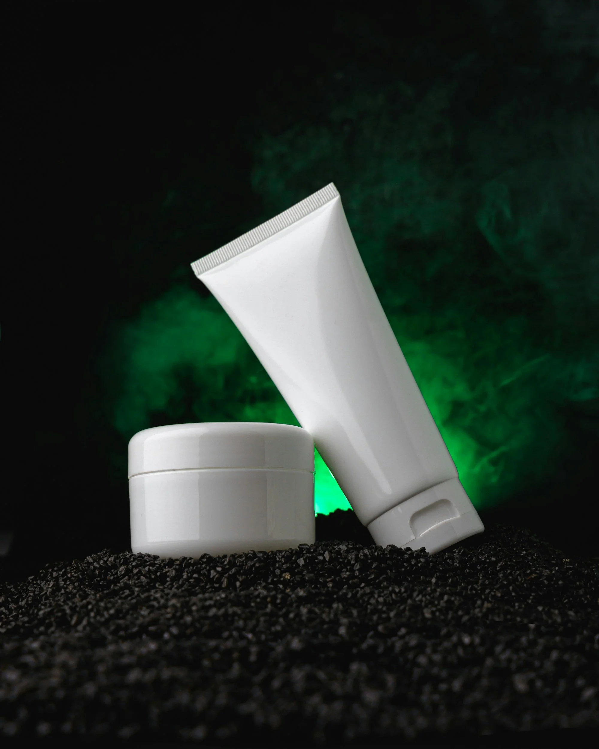White cosmetic tubes and lid on black granular surface with green smoke background.