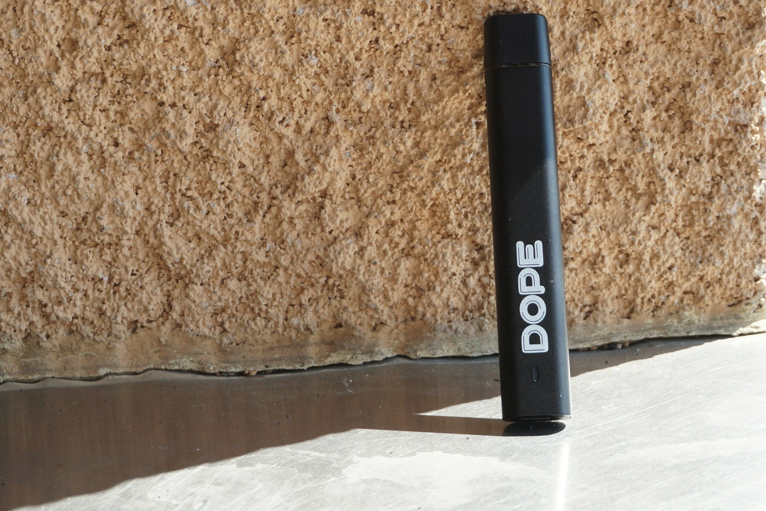 A black Dobe vape pen standing upright on a light-colored surface next to a textured brown wall.