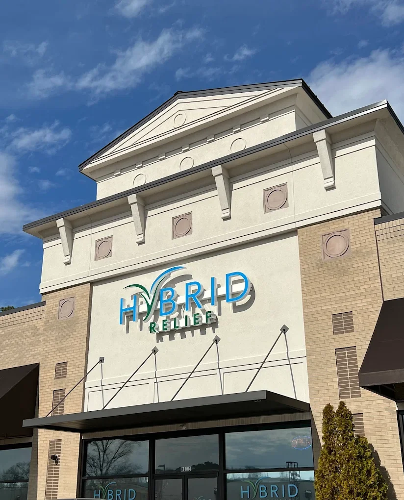 A building with a sign that says 'HYBRID RELIEF' in blue and green letters, with a leaf and wind symbol above the word 'HYBRID', and a clear blue sky in the background.