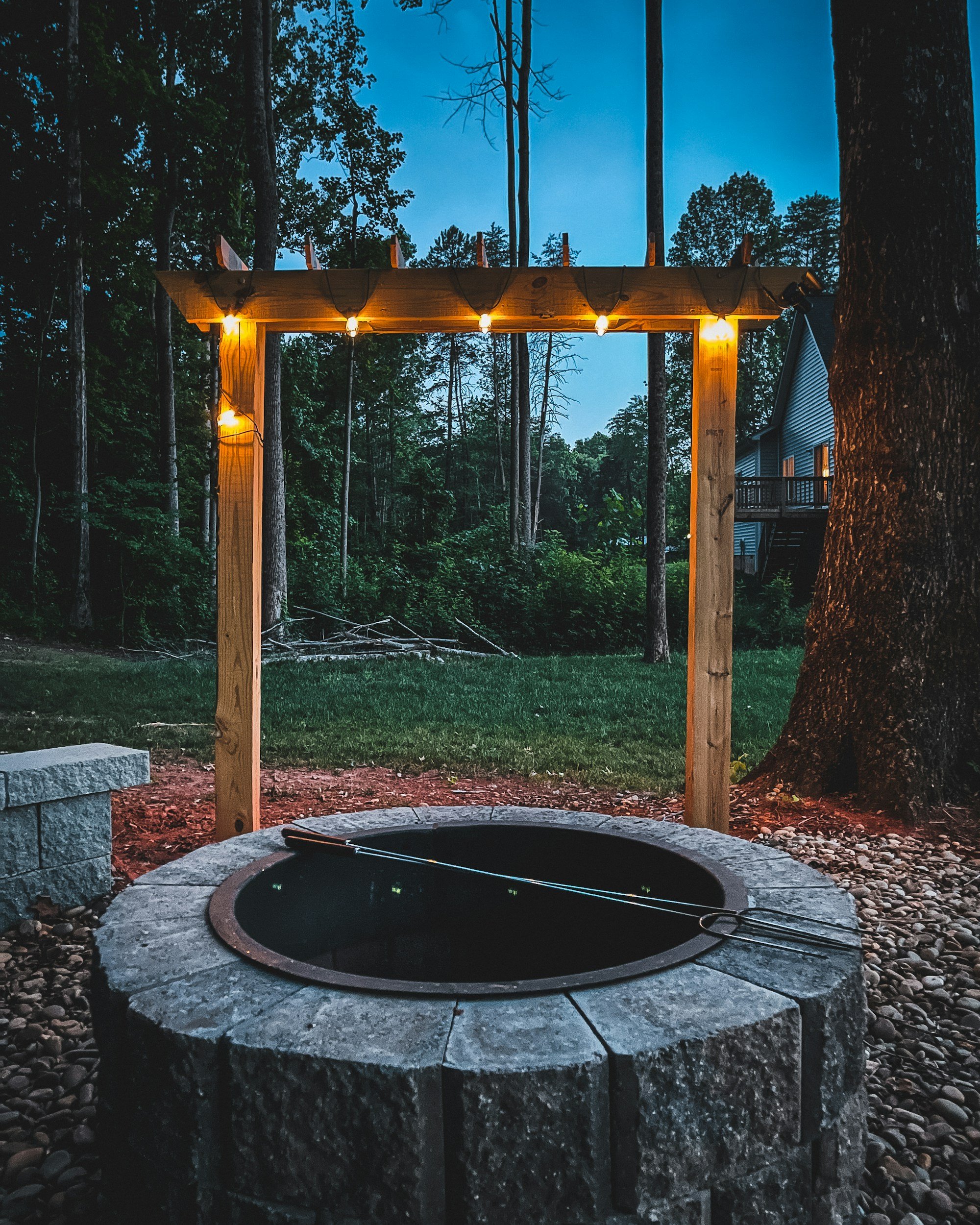 A fire pit surrounded by a stone border, with a swinging poker resting on its edge, outdoors at dusk. Behind it, there is a wooden arch with string lights, set in a wooded yard near a house.
