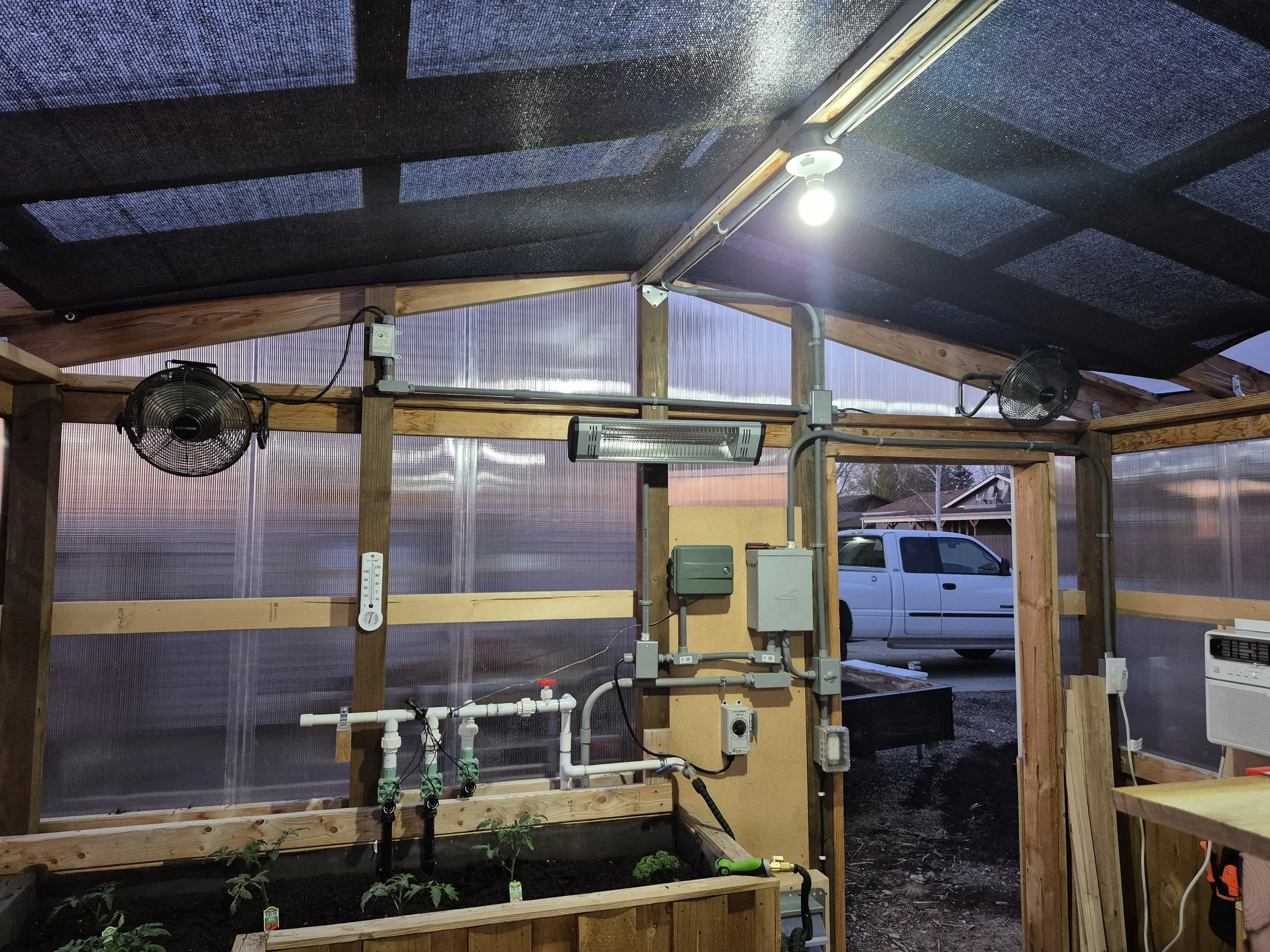 Inside a greenhouse with plants and gardening equipment, including fans, a heater, lighting, and sprinkler system during evening sunset.