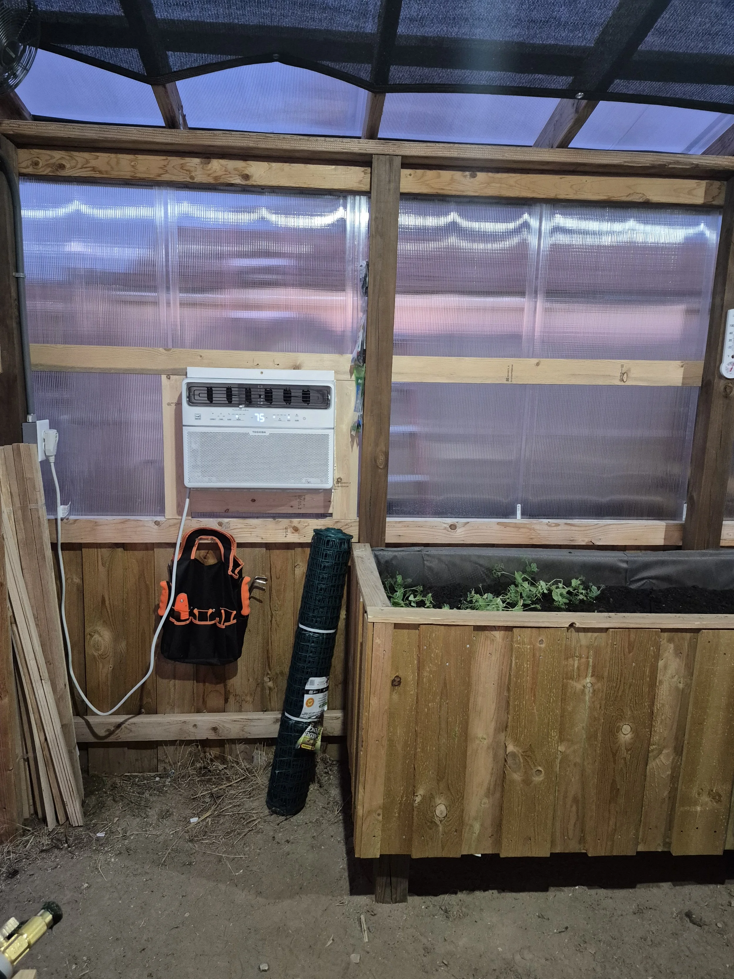 Inside a small greenhouse with wooden framing, plastic walls, and a sloped roof, a window air conditioning unit, a black and orange tool bag hanging on the wall, and a rolled-up black garden mesh are visible. There is a raised wooden planter with plants growing inside.