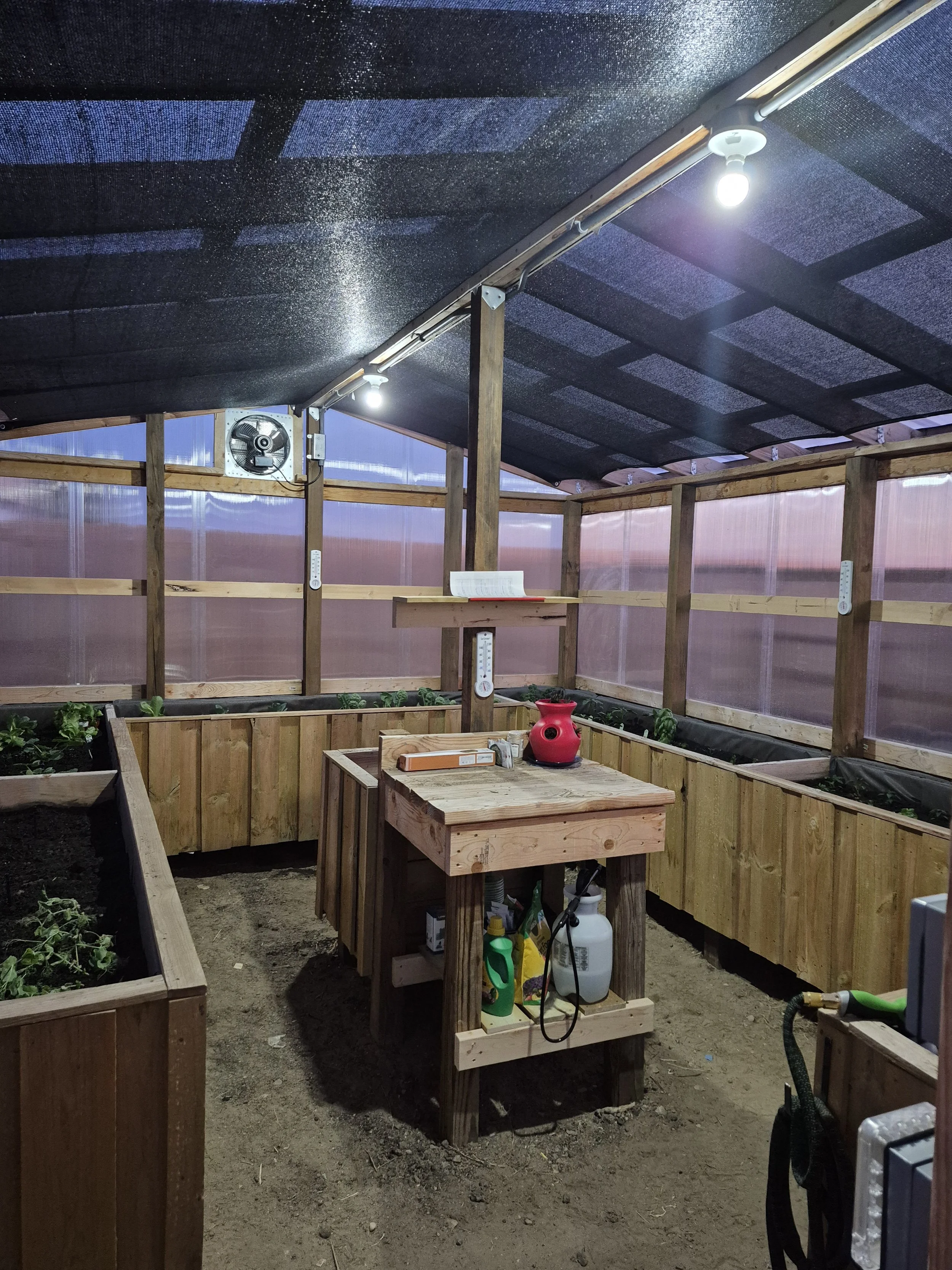 A small indoor garden or greenhouse with wooden raised beds for plants, a wooden work table with gardening supplies, and a fan on the wall, all under a black ceiling with bright lighting.