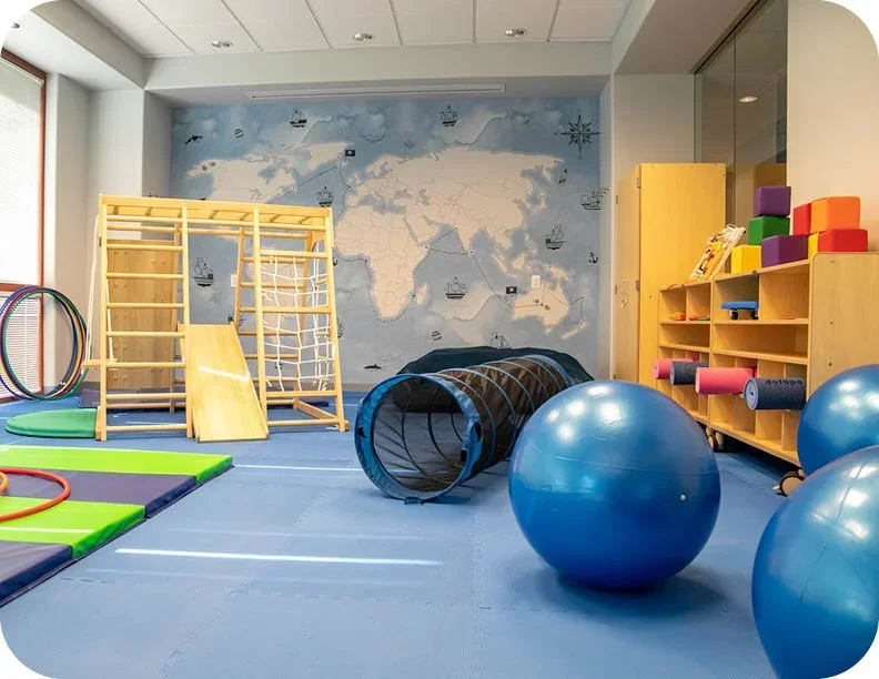 Children's indoor playroom with a blue world map mural on the wall, wooden climbing structure, colorful foam mats, hoops, blue exercise balls, and shelves with foam rollers and toys.