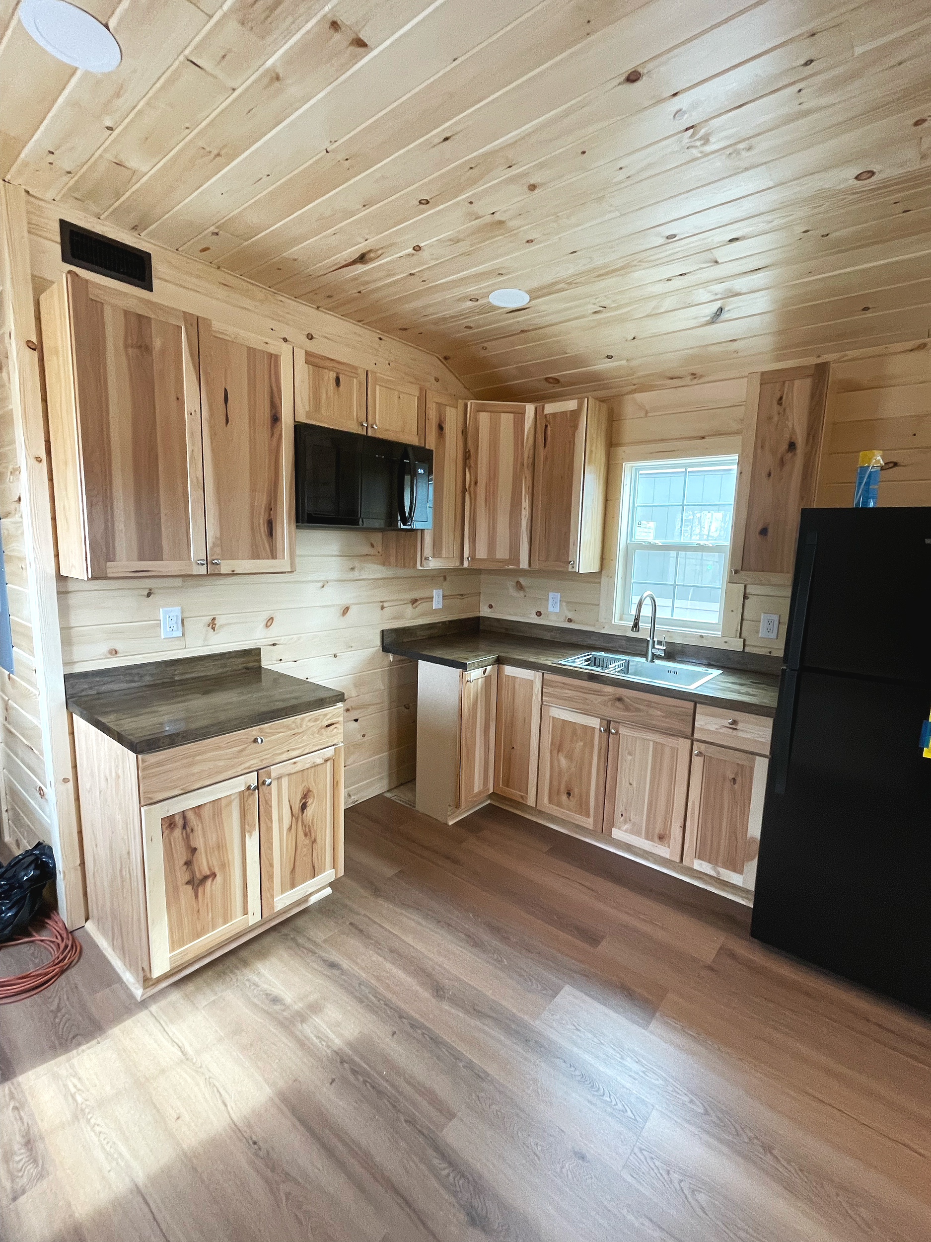 A kitchen with wood paneling on the walls and cabinets, a black refrigerator, a stainless steel sink, an oven, and a window above the sink.