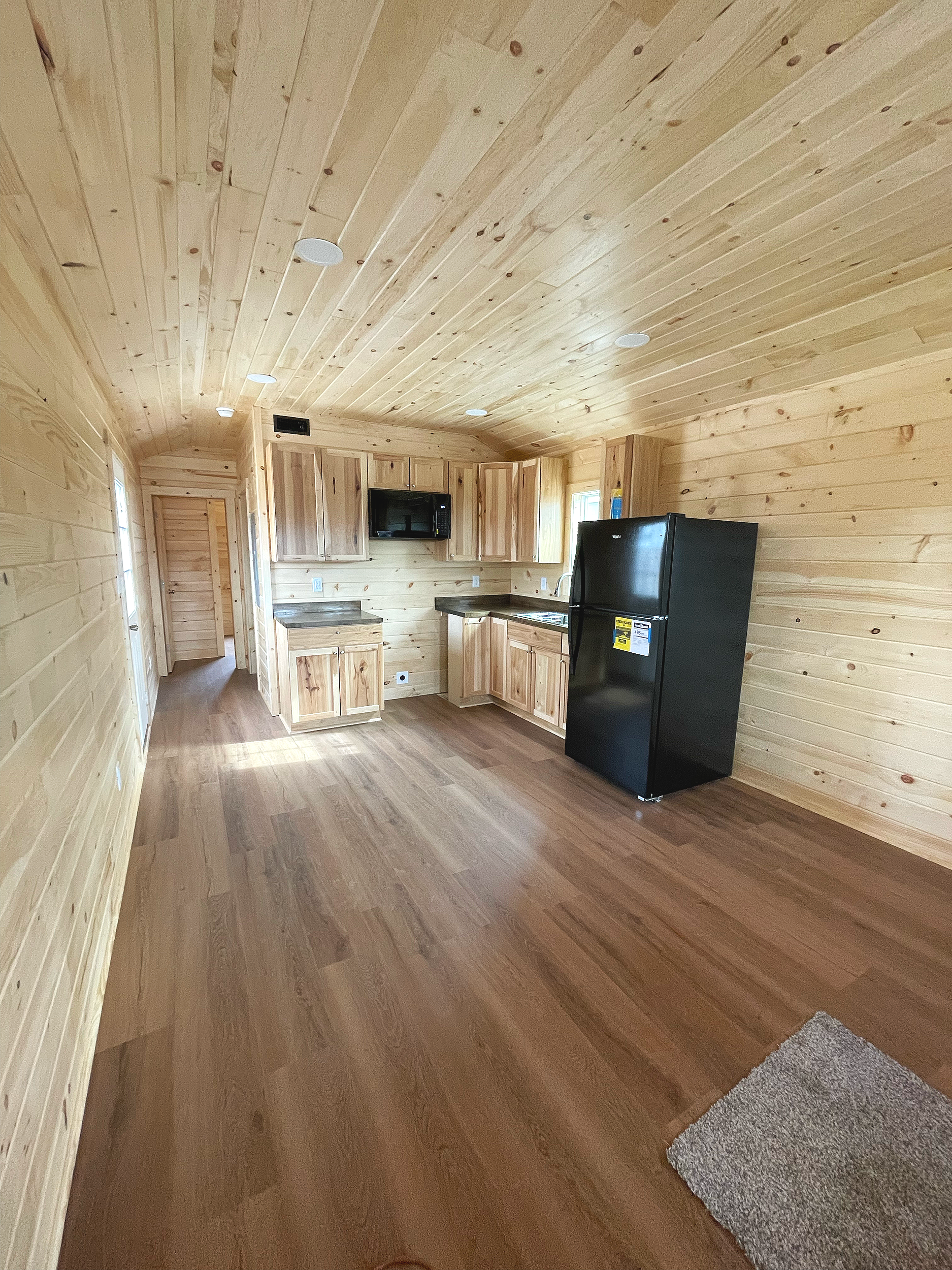 Interior of a wooden cabin with wood-paneled walls and ceiling, hardwood floor, a black mini refrigerator, wooden kitchen cabinets, a window, a small countertop, along with a rug on the floor.