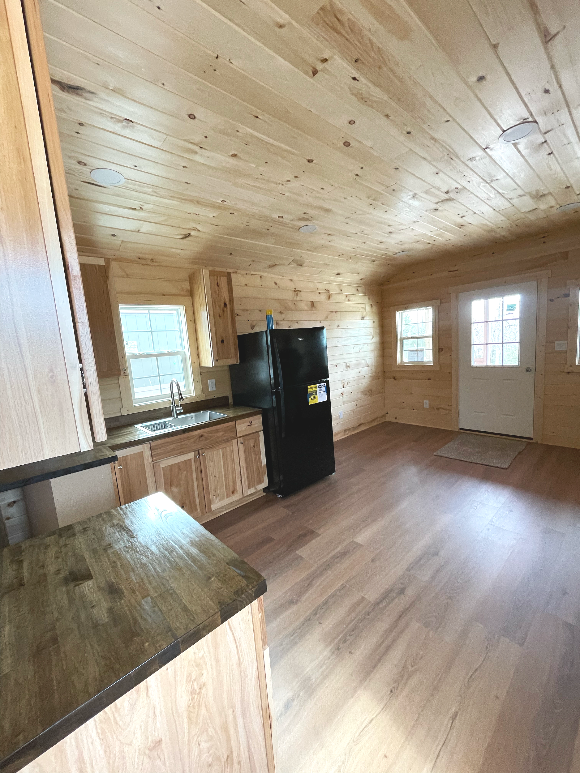 Interior view of a wooden cabin with a kitchen area and a door leading outside, natural light coming through windows, wood-paneled walls, and hardwood flooring.
