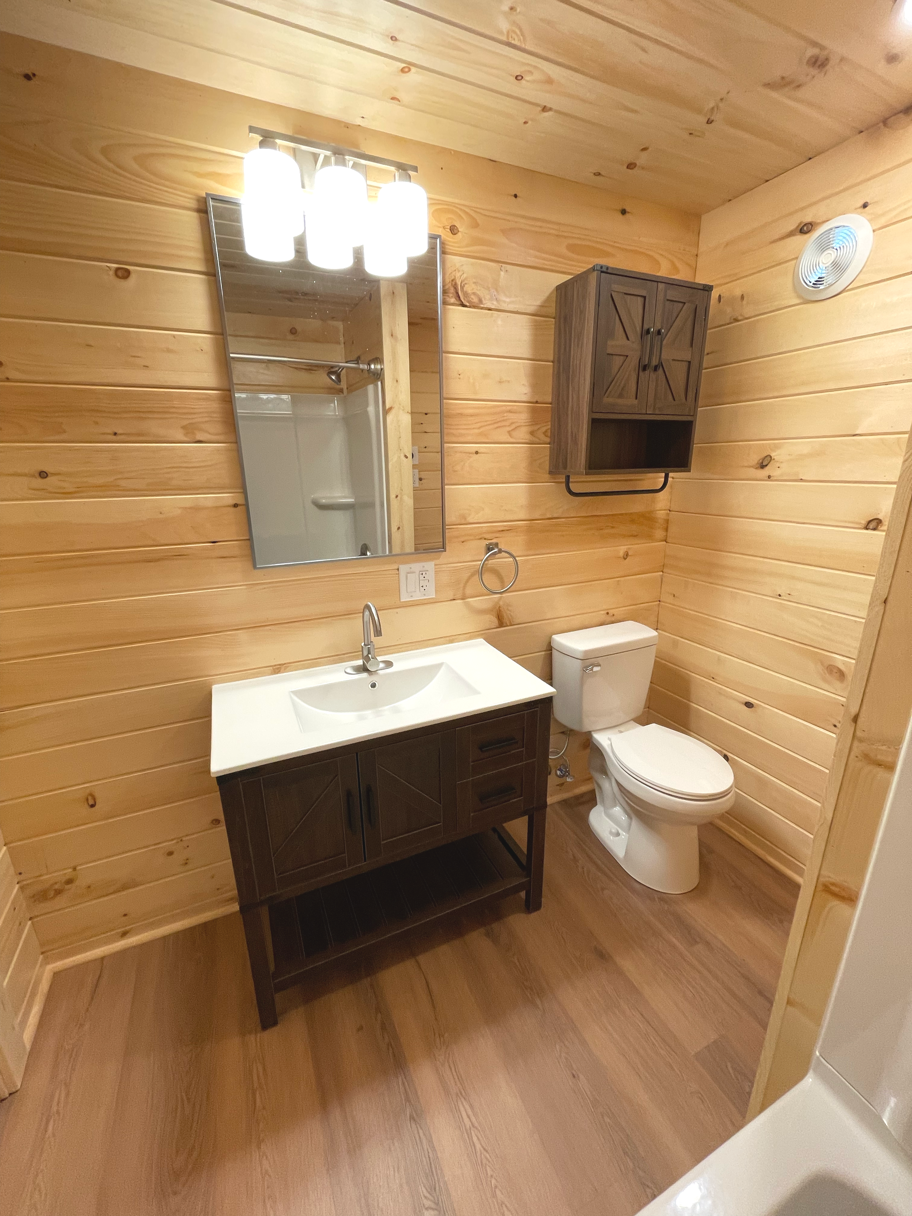 A wooden bathroom with natural wood walls and flooring, featuring a white toilet, a dark wooden cabinet, a mirror, and a bathroom sink with a faucet. There is a light fixture with multiple bulbs above the mirror and a small vent on the ceiling.