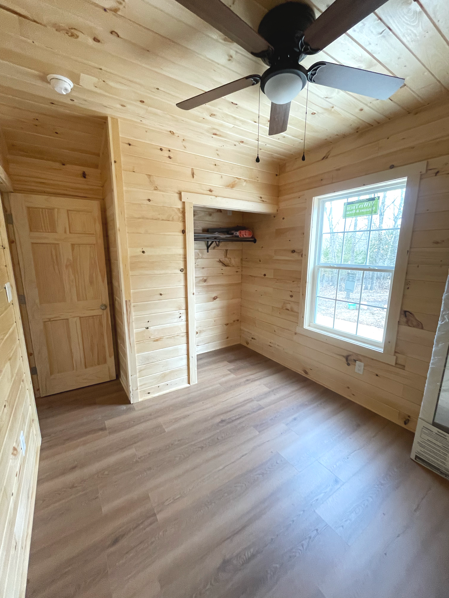 Interior view of a room with wooden walls and floor, a window showing trees outside, and a wooden door, with some items like a fan and a disassembled table.