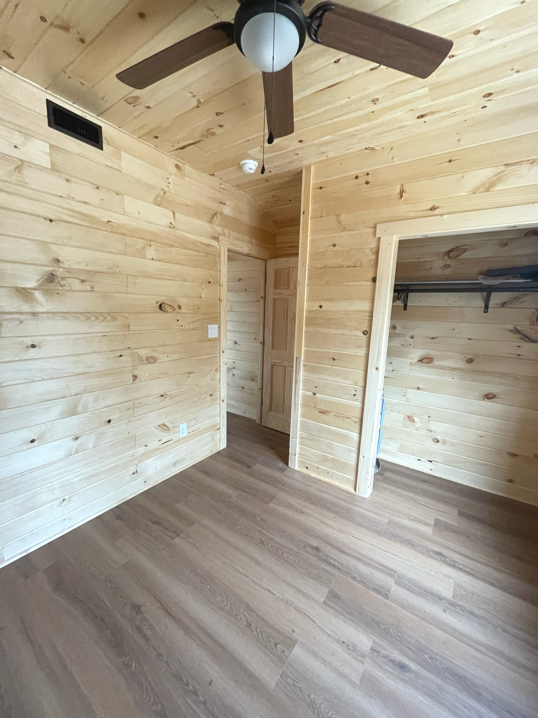 A room with wooden walls and flooring, featuring a large mirror on the wall, a window, and a ceiling light fixture.
