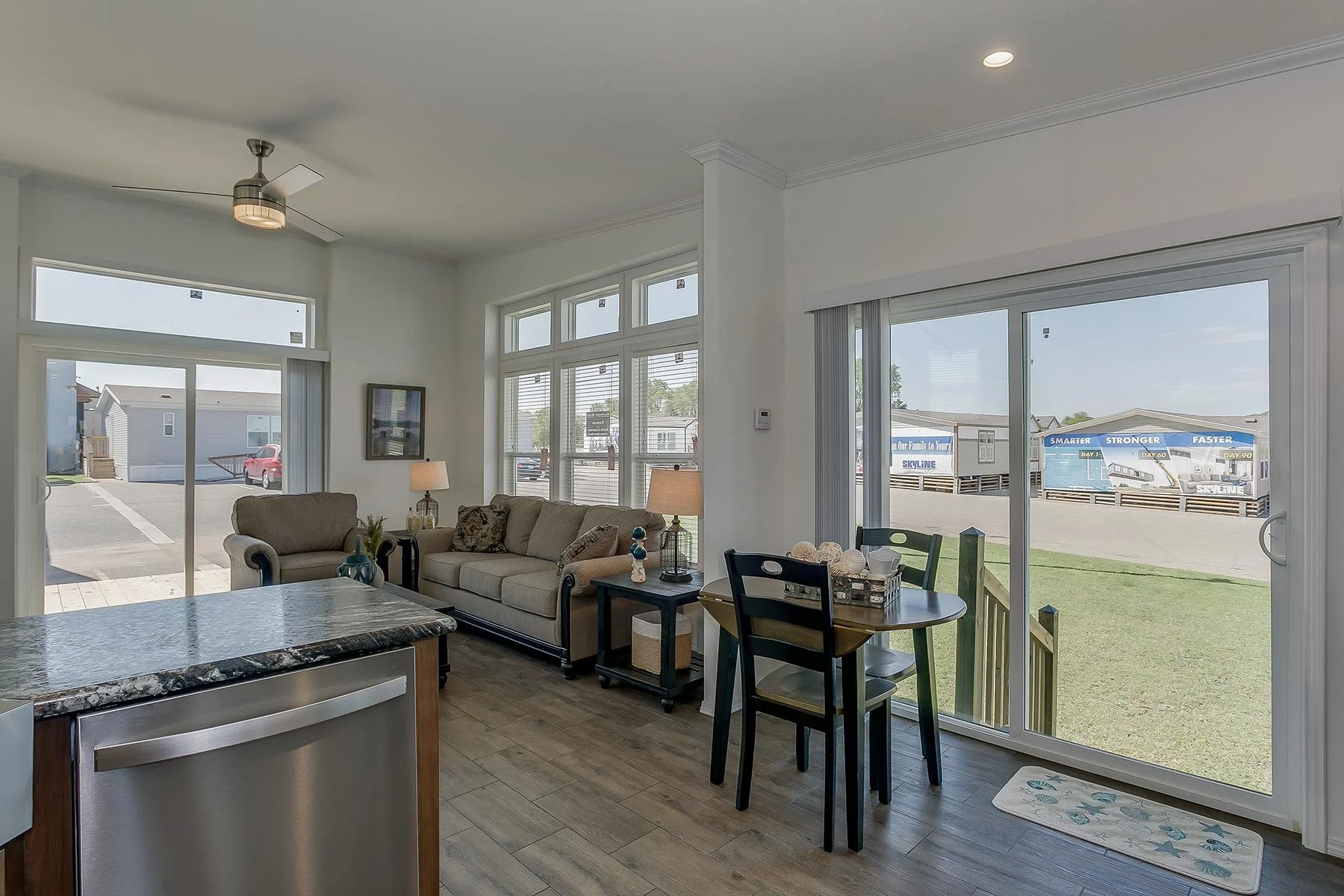 Living room with beige couches, side tables with lamps, window blinds, sliding glass door, and outdoor view of a parking lot and neighboring houses.
