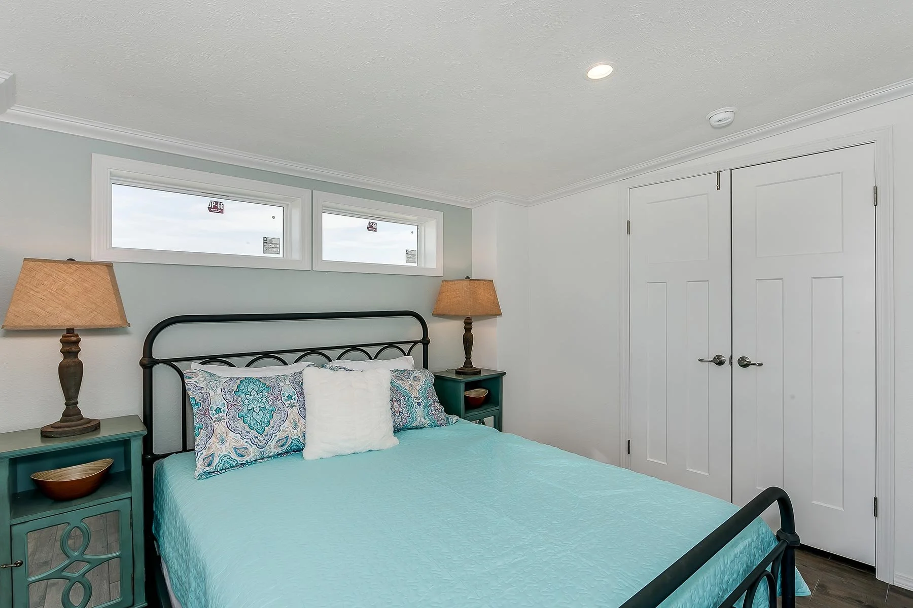 A bedroom with a bed, two nightstands with lamps, and a closet with double doors. The bed has a black metal frame, a light blue quilt, patterned pillows, and a white decorative pillow. There are three small horizontal windows above the bed, with a wh