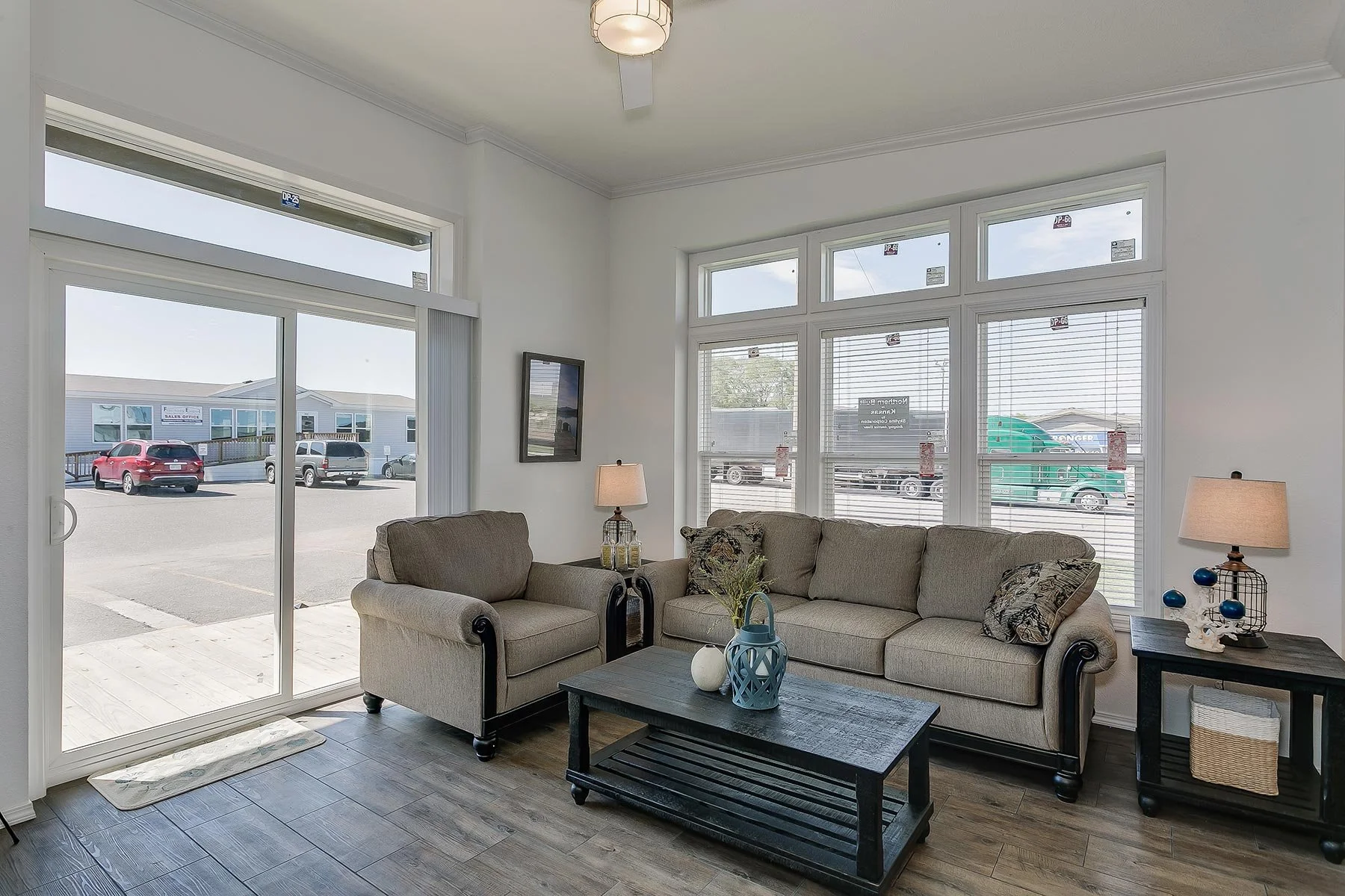 Living room with beige couch, wooden coffee table, side table with lamp, and large windows letting in natural light, overlooking parking lot with cars and commercial buildings.