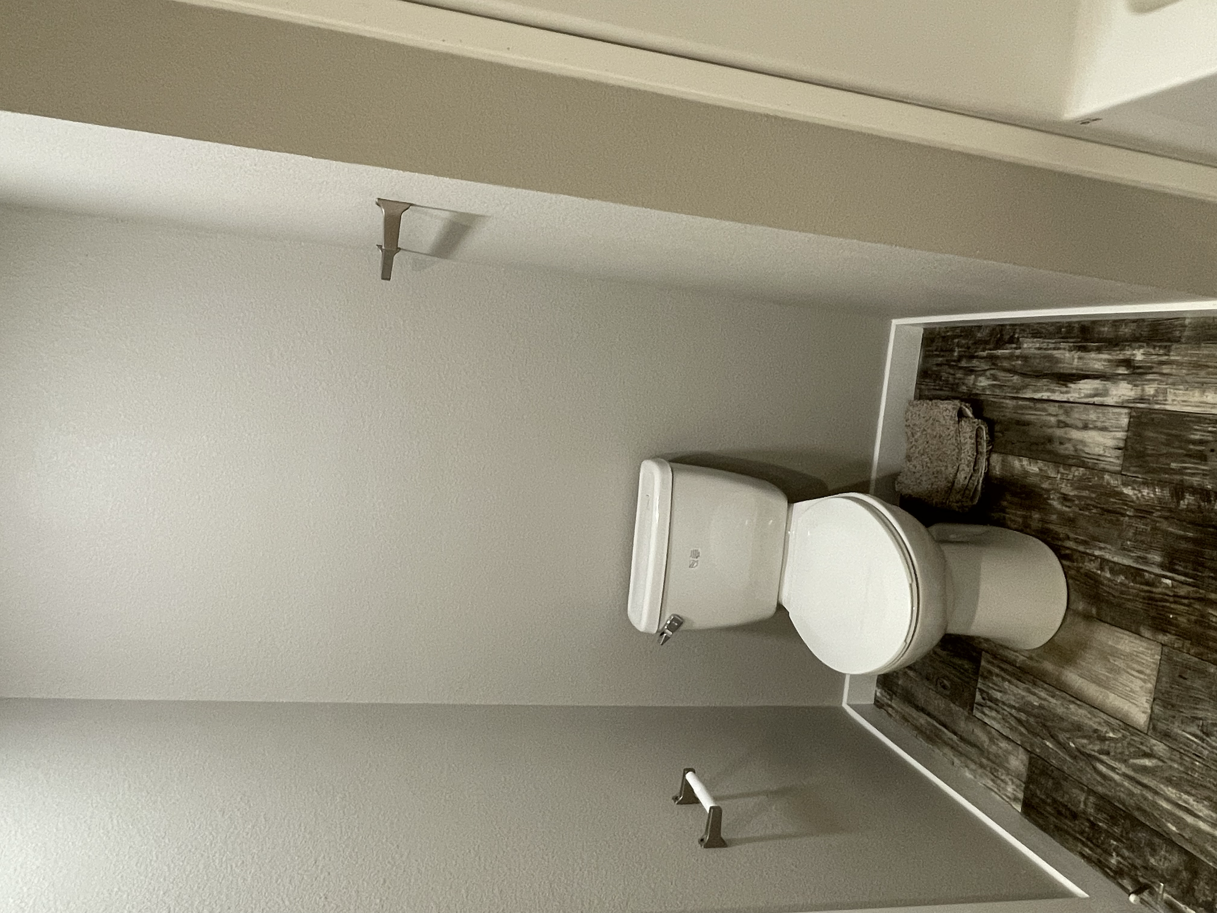 A bathroom with a white toilet and patterned wooden floor. The wall above the toilet is plain, with an unfinished wall fixture or pipe hanging from it.