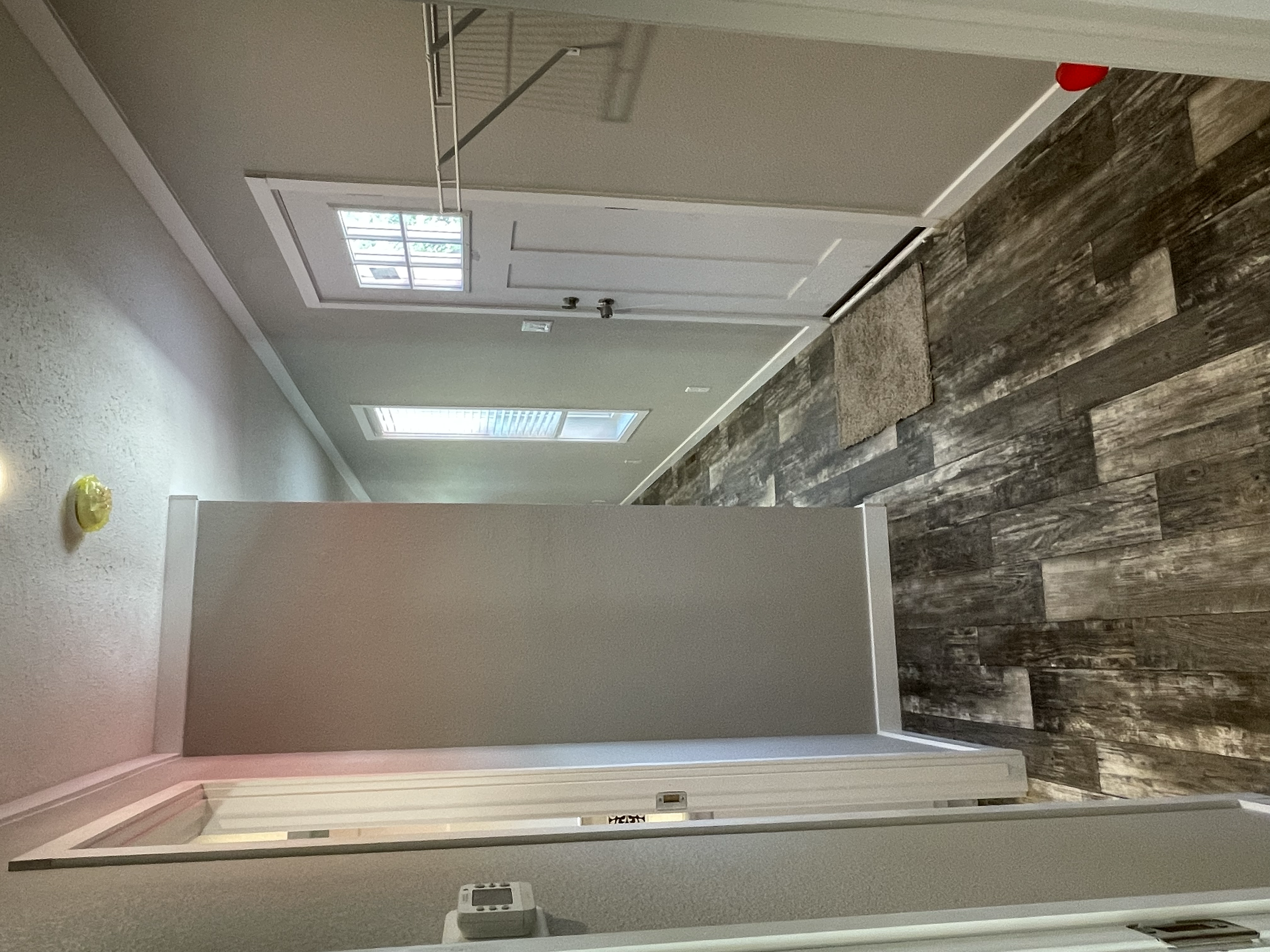 An interior hallway with gray and brown wood-look flooring, beige walls, a white door, windows with white blinds, and a red object near the ceiling.