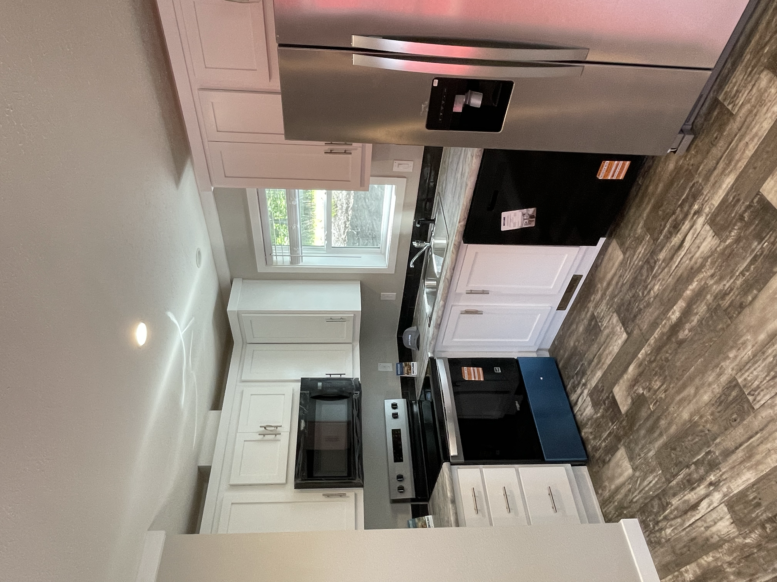 Modern kitchen with white cabinets, black appliances, a stainless steel refrigerator, and a window above the sink showing greenery outside.