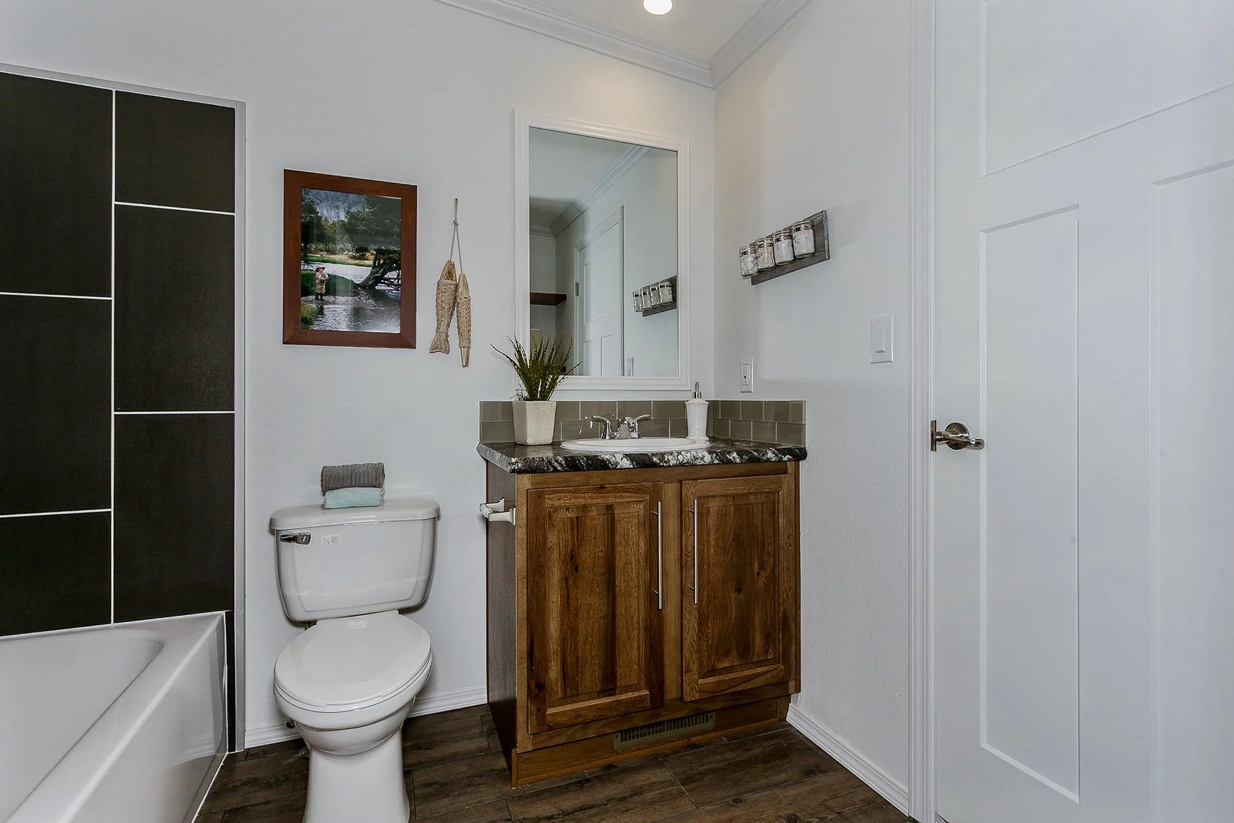 A modern bathroom with a white toilet, a wooden vanity with a dark marble countertop, and a large mirror. There is a framed picture of a outdoor scene hanging on the wall, along with some decorative fish hanging beside it. The bathroom has white wall