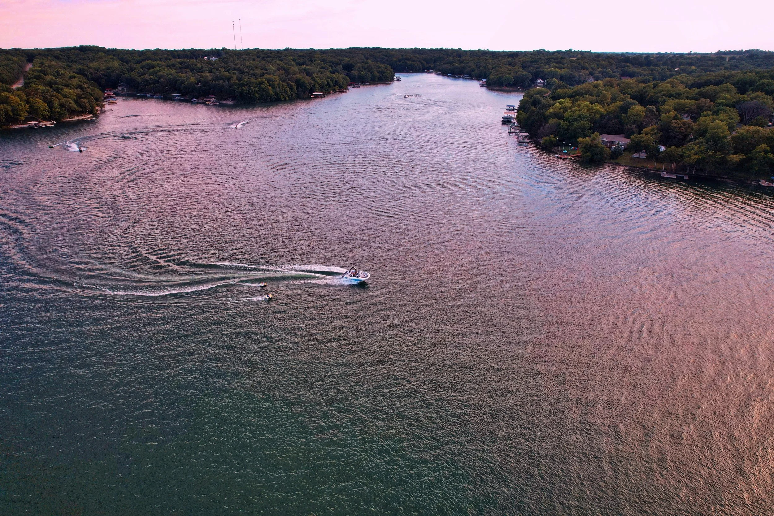 Aerial view of a wide river with boats creating wake on the water, surrounded by tree-lined shores and houses with docks during sunset.